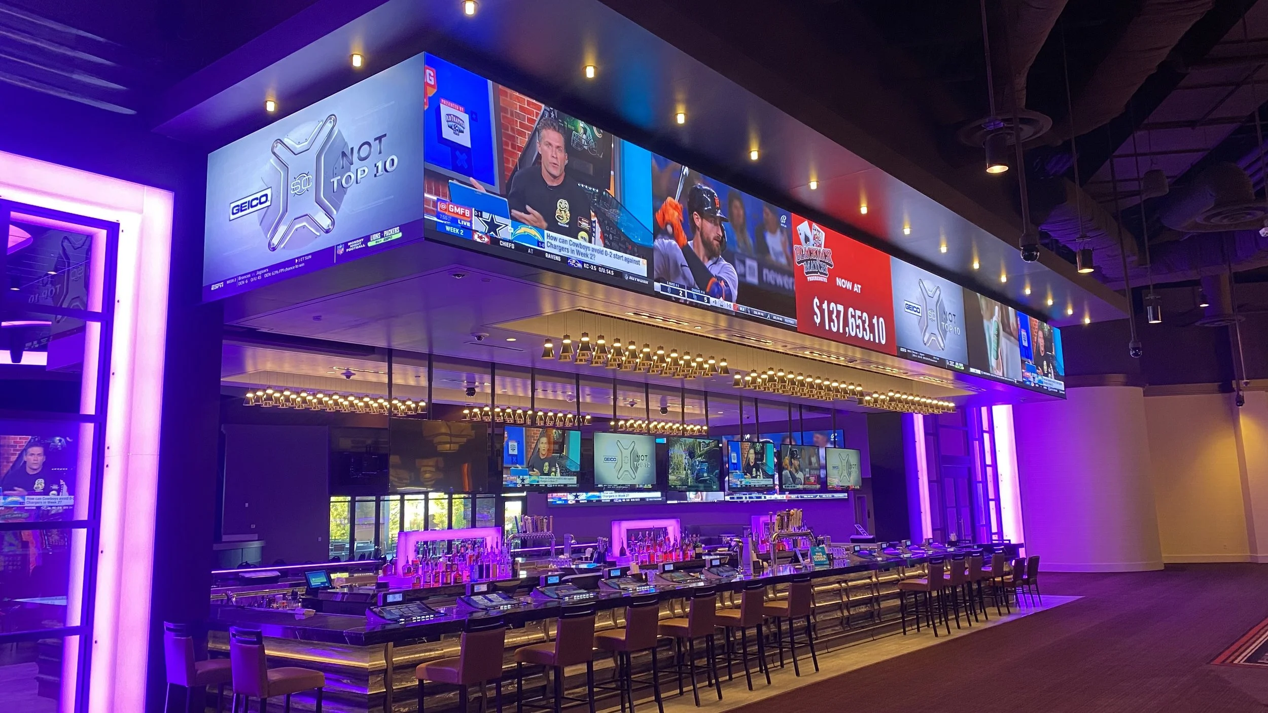 An empty sports bar or casino interior with purple lighting, multiple television screens showing sports and betting advertisements, a row of electronic gaming or betting terminals with chairs, and a bar area with bottles and glasses illuminated by pink and purple lights.