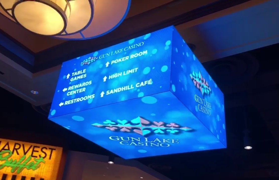 Blue illuminated cube sign with white text indicating directions to Gun Lake Casino amenities, including poker room, table games, rewards center, restrooms, high limit, and Sandhill Café, with some arrows pointing up and others left.