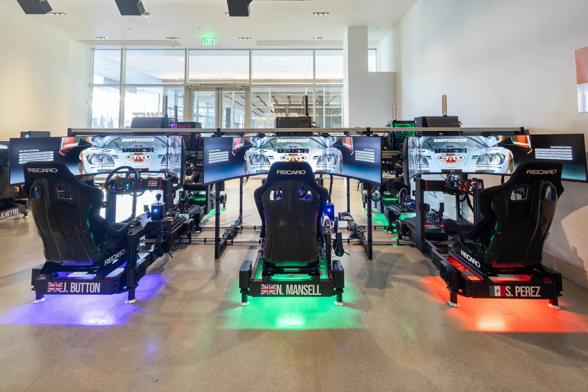 Three racing simulator setups with multiple monitors, racing seats, and simulation controls inside a brightly lit room with large glass doors and an exit sign.