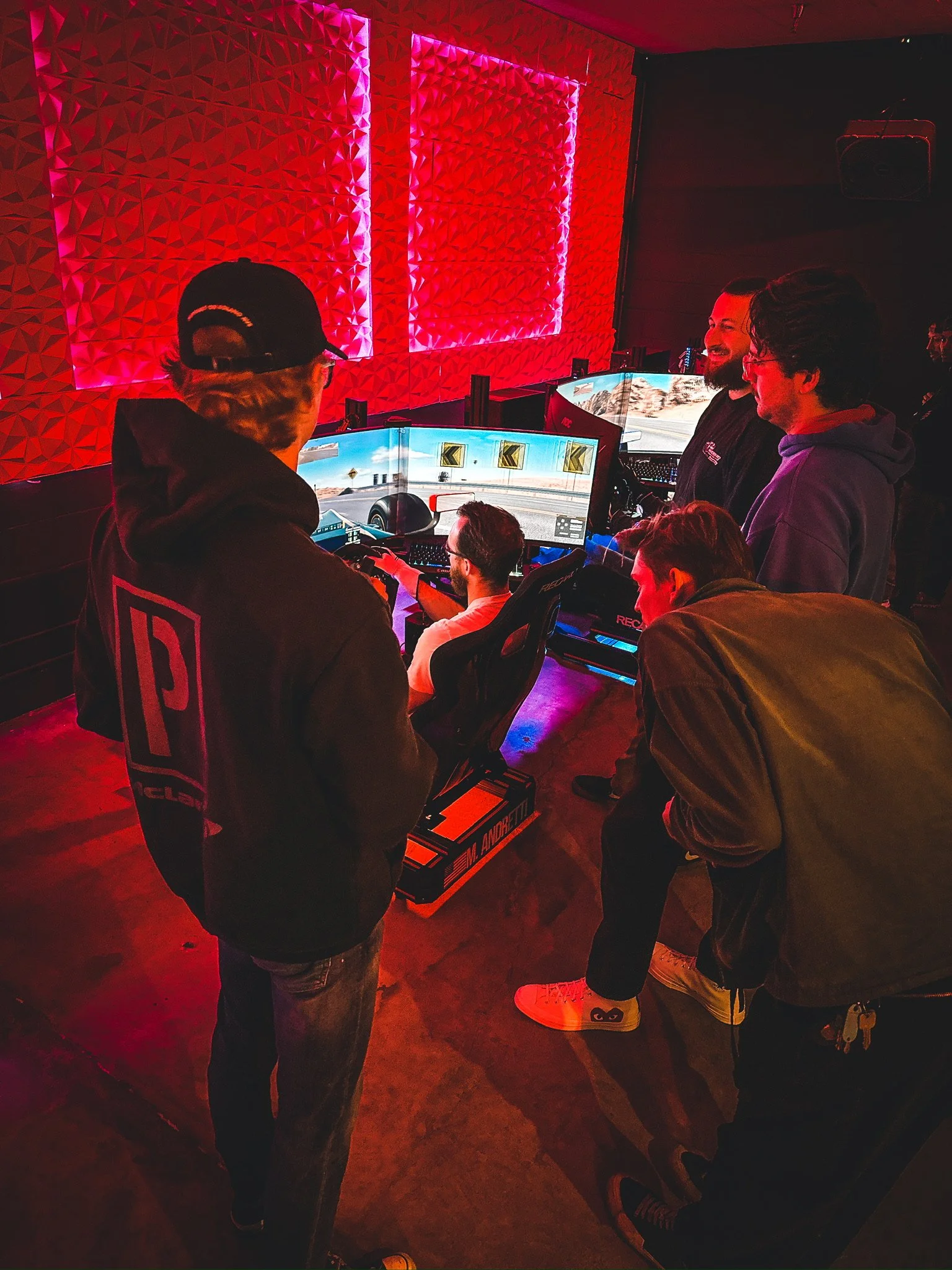 A group of people watching and participating in a racing simulation game session. The room is dimly lit with red ambient lighting and textured walls. Several computer monitors display a racing track scene. One person is seated in a racing simulator setup with a wheel and pedals, while others stand nearby, observing and discussing.