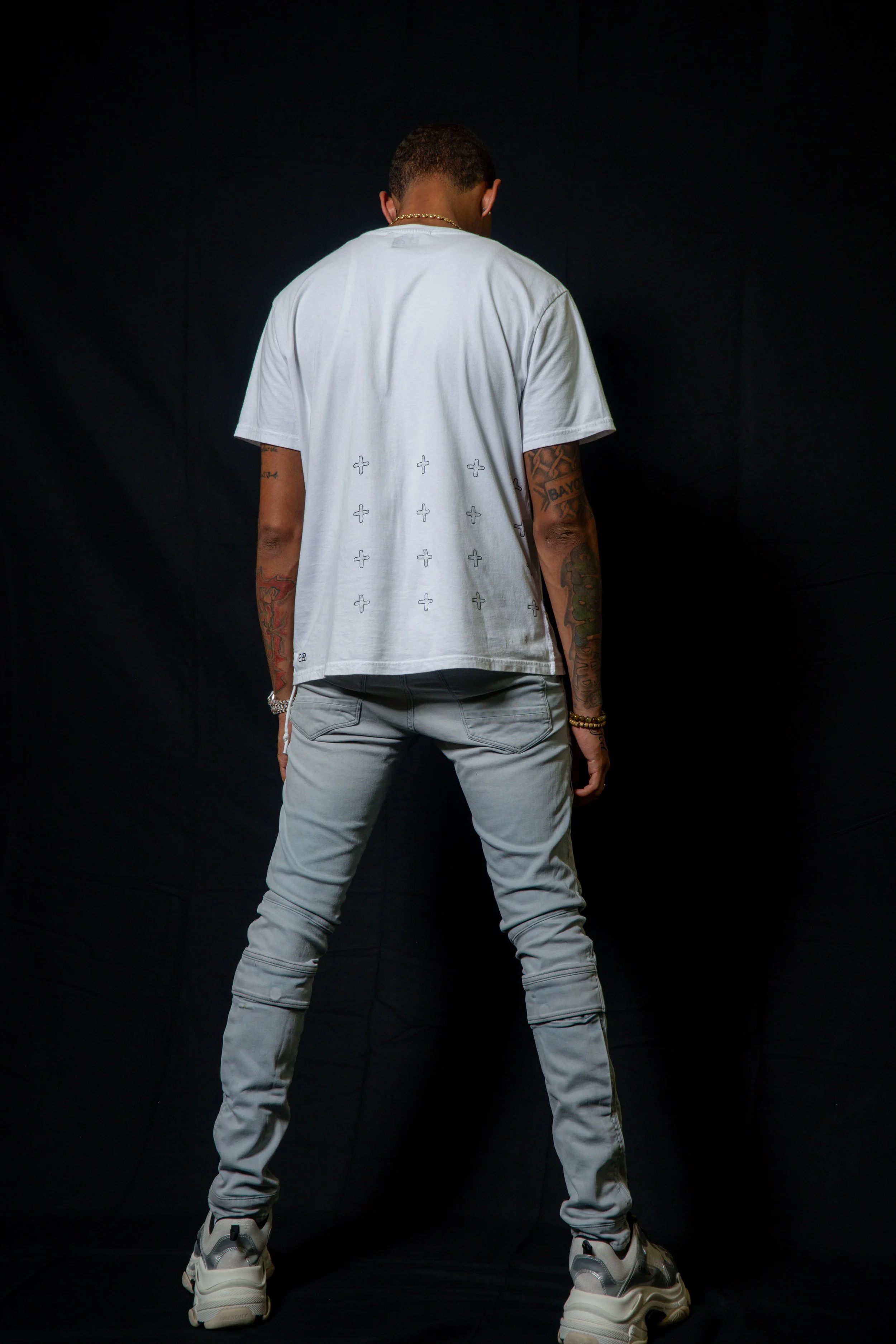 Rear view of a man standing with legs apart against a black background, wearing a white T-shirt, light gray jeans, and sneakers, with tattoos on his arms.