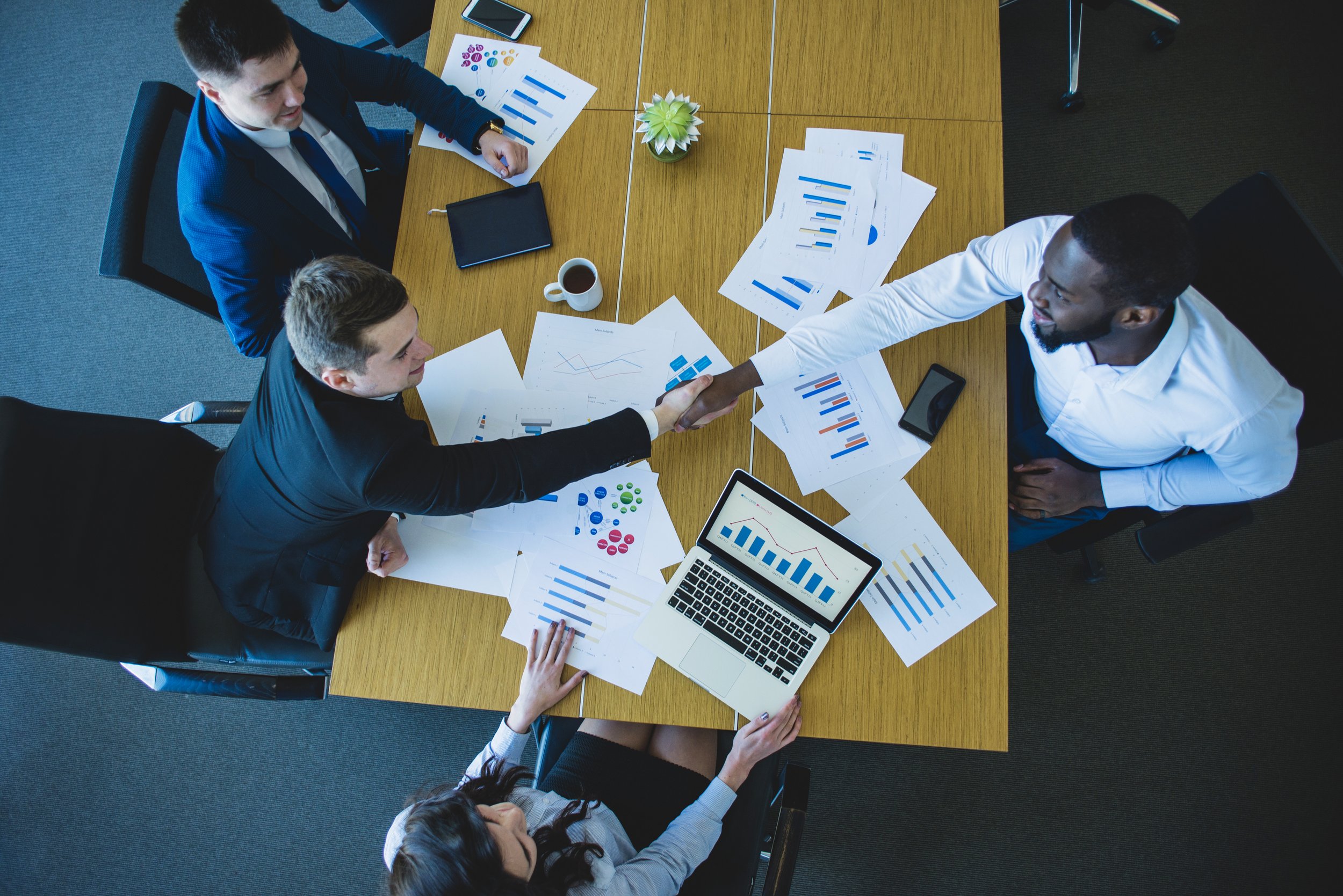 Business professionals shaking hands over a conference table filled with charts, graphs, a laptop, and documents in a meeting room.