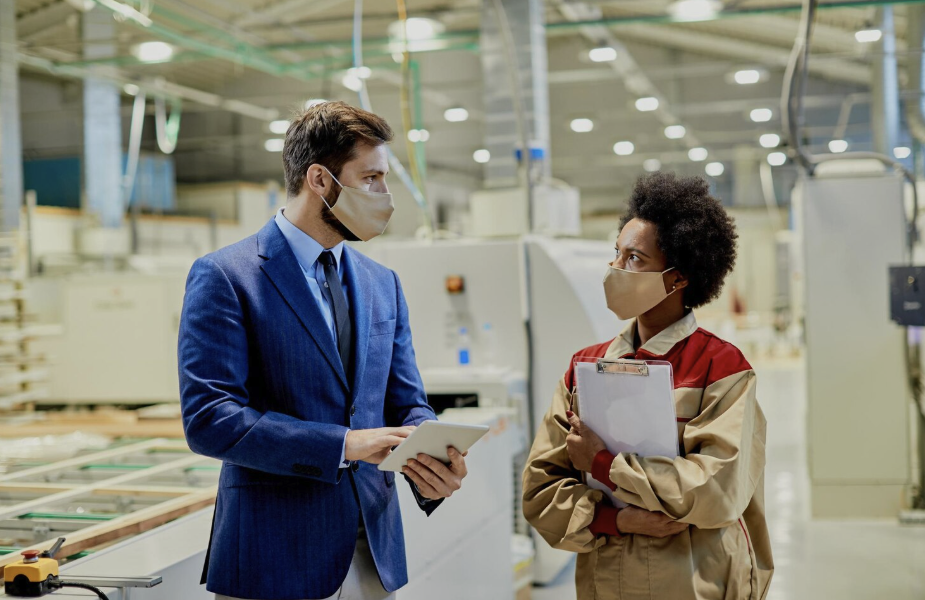 Two factory workers, a man in a suit and a woman in work uniform, wearing face masks and having a conversation in an industrial manufacturing setting.