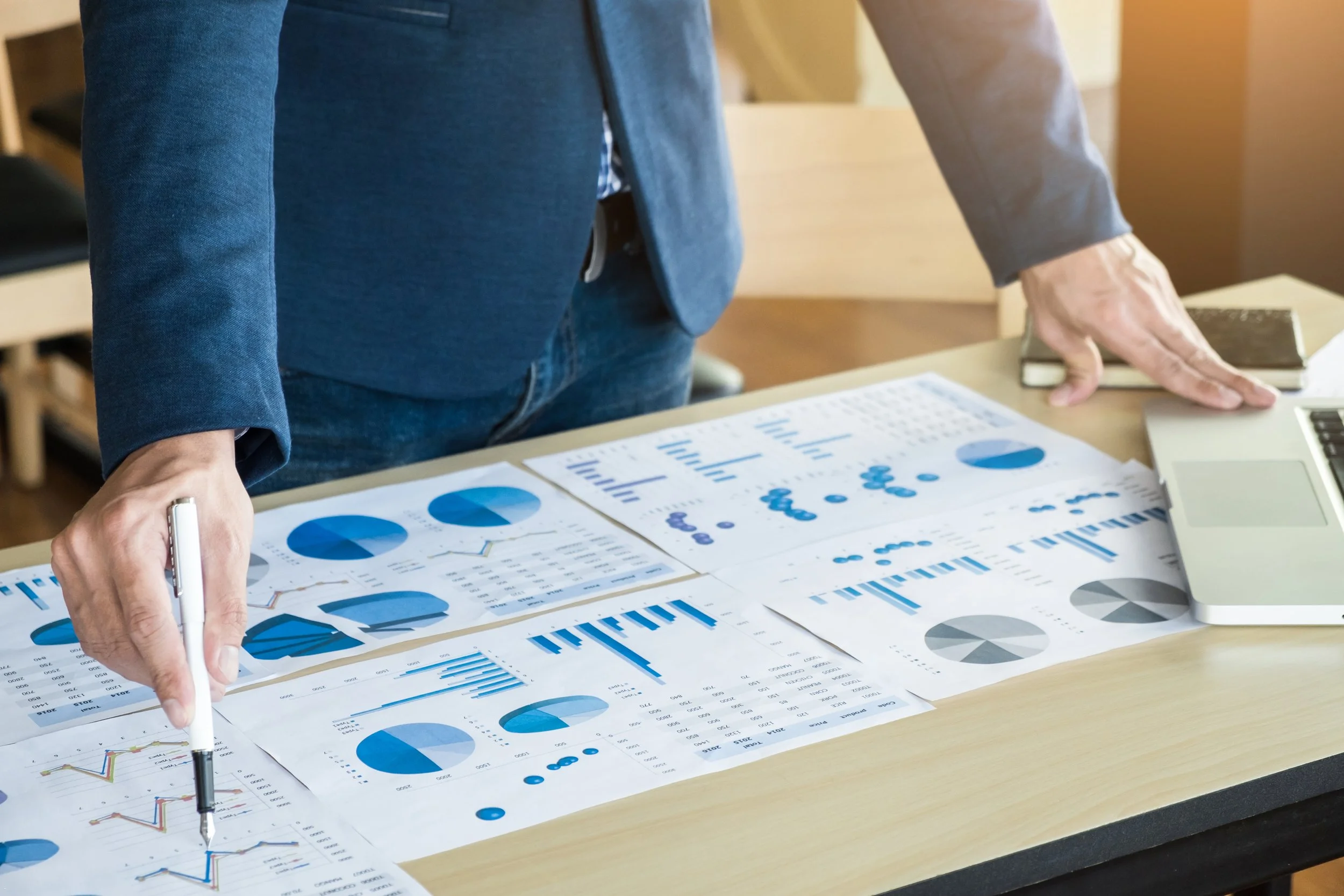 Person in a blazer analyzing blue and gray pie charts, bar graphs, and line graphs on a desk, using a pen and a laptop.