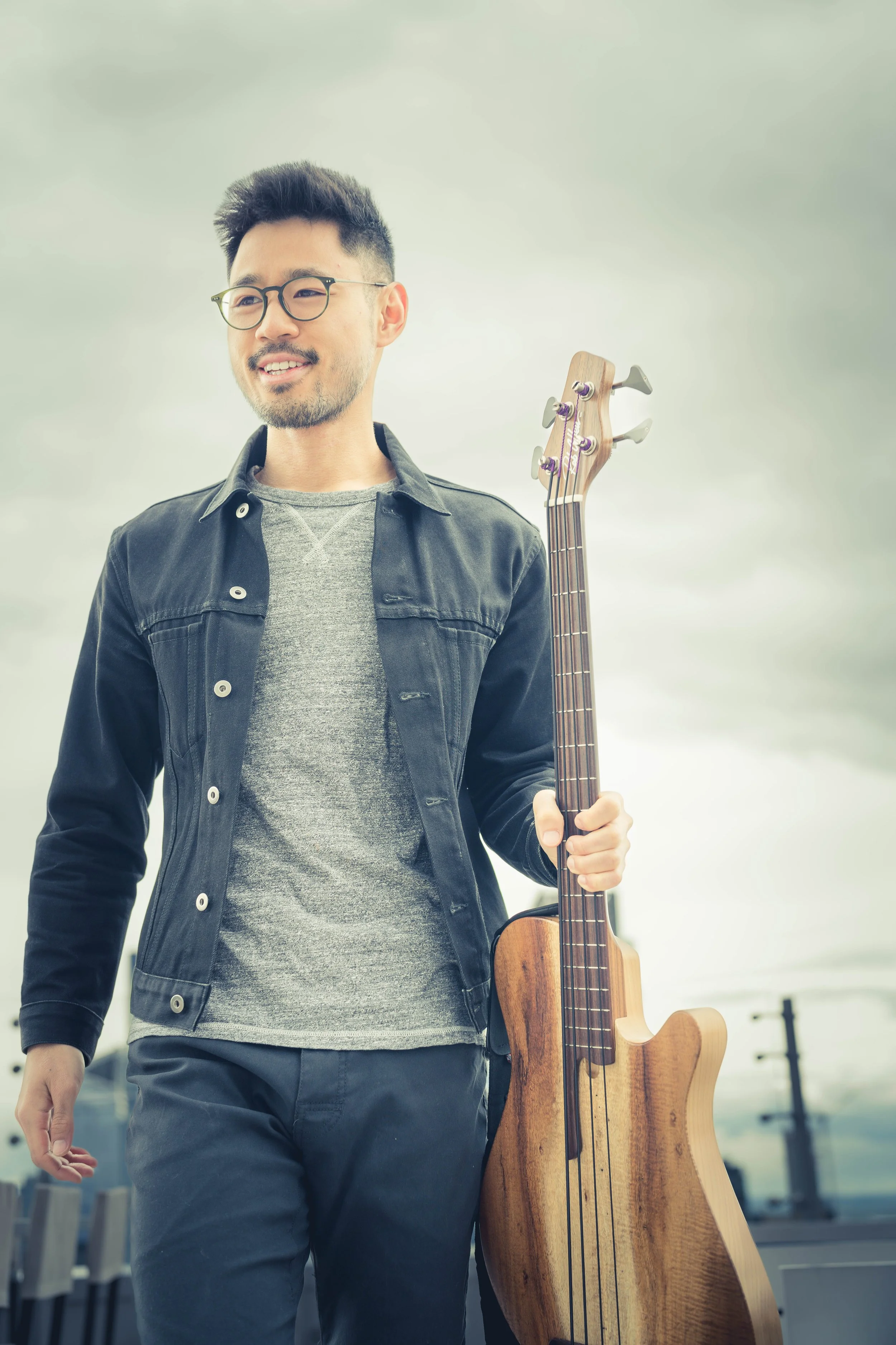 A man wearing glasses, a black denim jacket over a gray shirt, holding a wooden electric bass guitar, outdoors on a cloudy day.