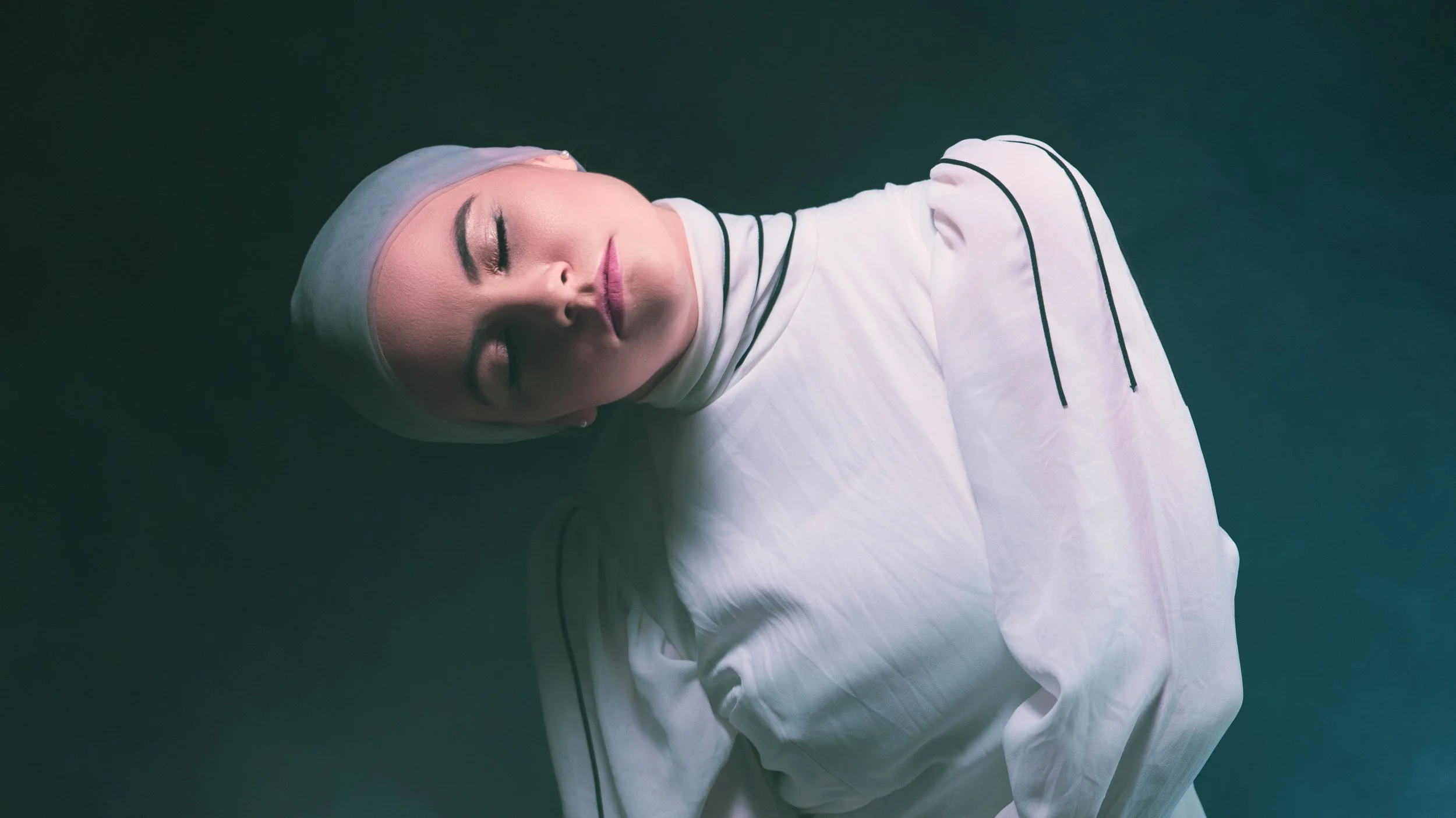 A cinematic portrait of a woman in a white high-neck dress and light grey headscarf, leaning her head back with closed eyes against a dark, moody background.
