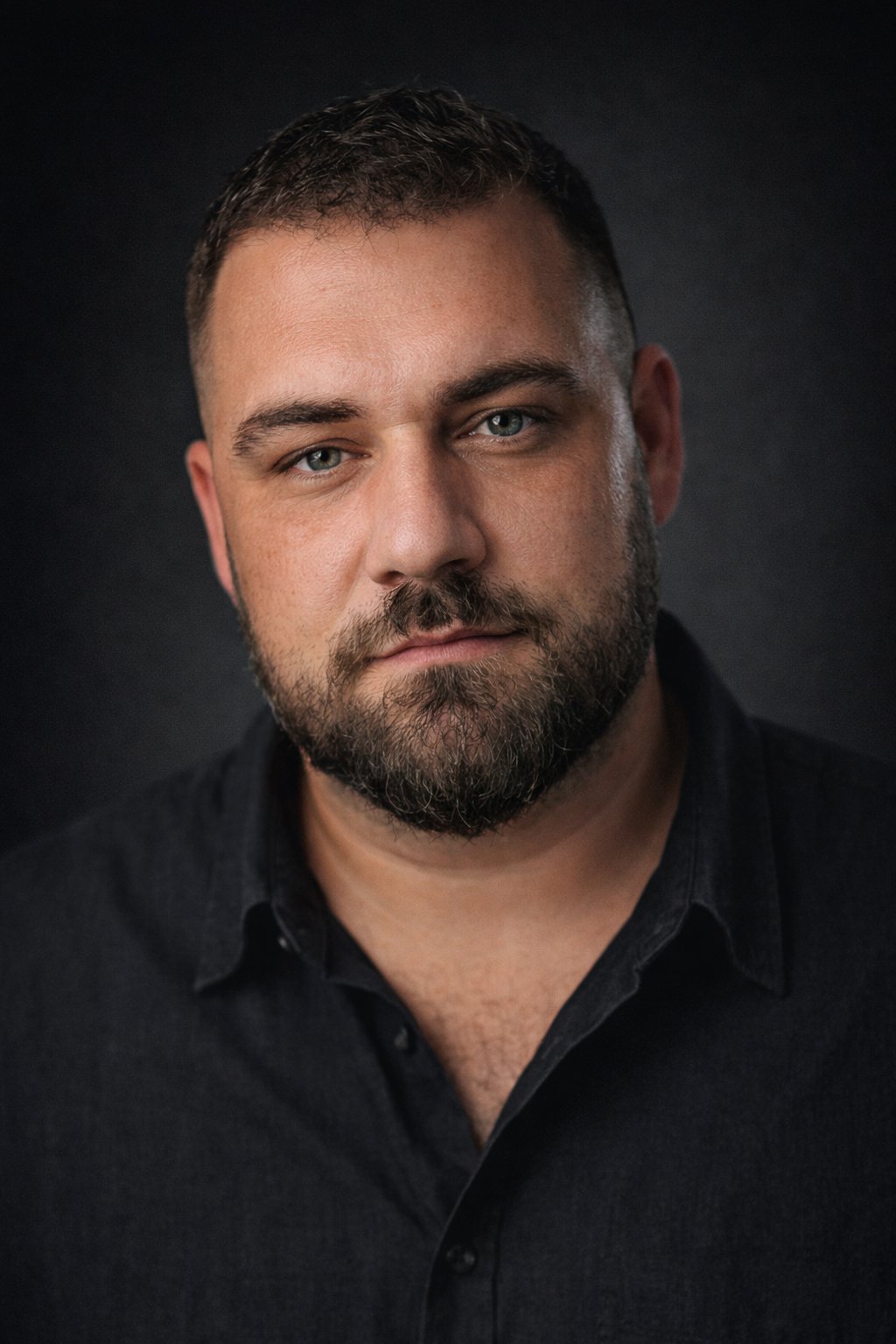 Close-up portrait of a man with short dark hair, blue eyes, and a beard, wearing a black shirt against a dark background.