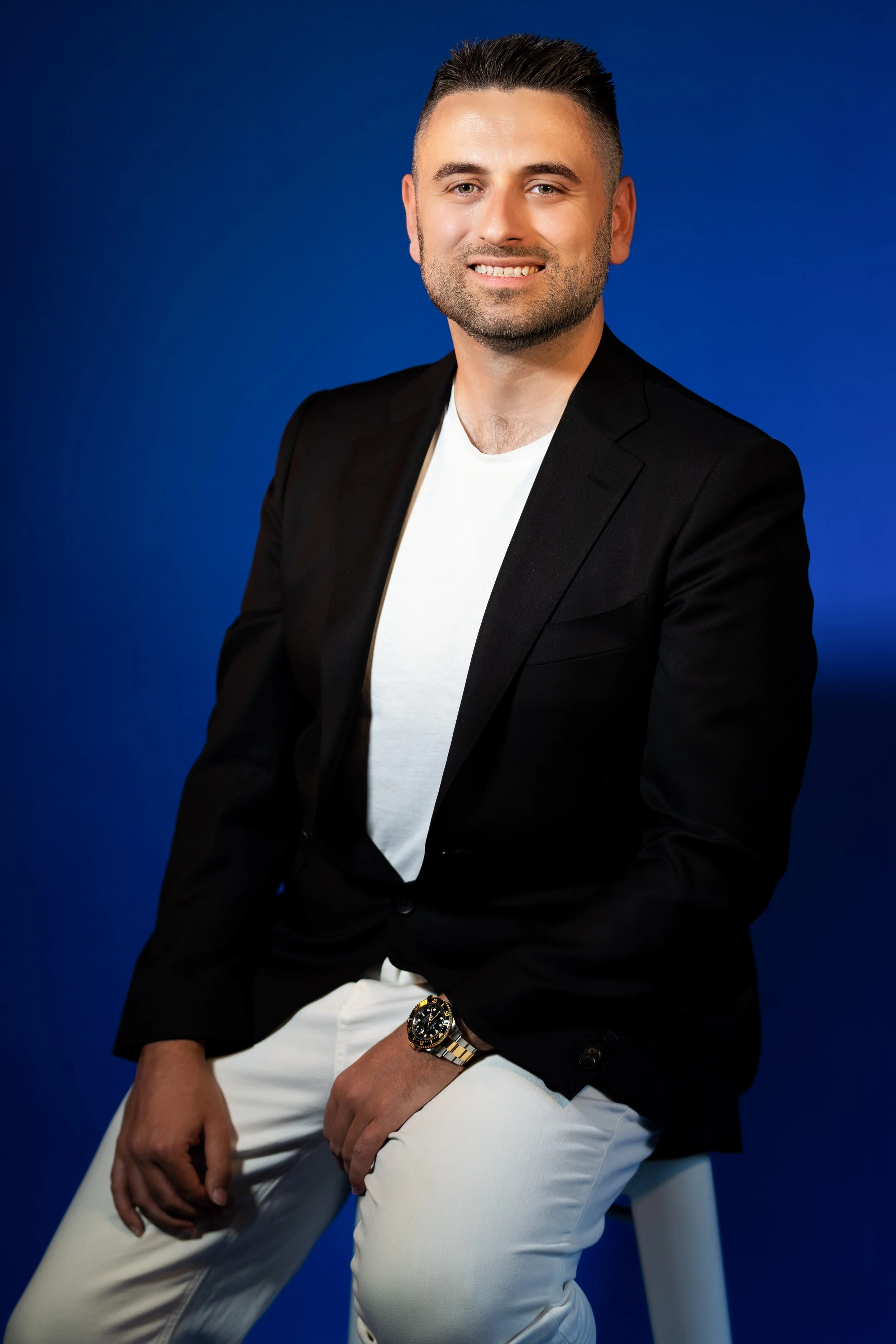 A young man with dark hair, beard, and light skin wearing a black blazer, white t-shirt, white pants, and a gold and silver watch, sitting against a deep blue background.