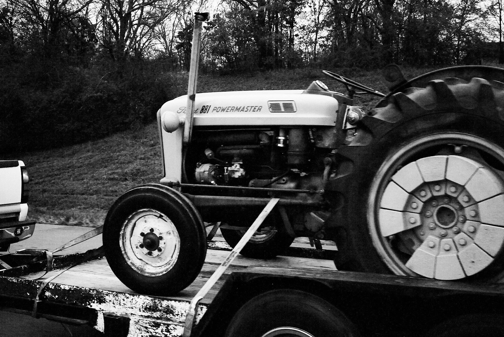 A vintage Ford 8N Powermaster tractor on a flatbed trailer, secured with straps, parked outdoors with trees and bushes in the background.