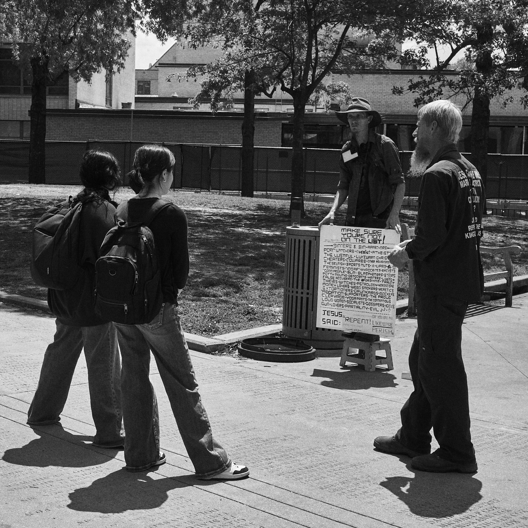 A man with a beard and a woman with gray hair holding a sign, speaking to two women with backpacks in a park on a sunny day.