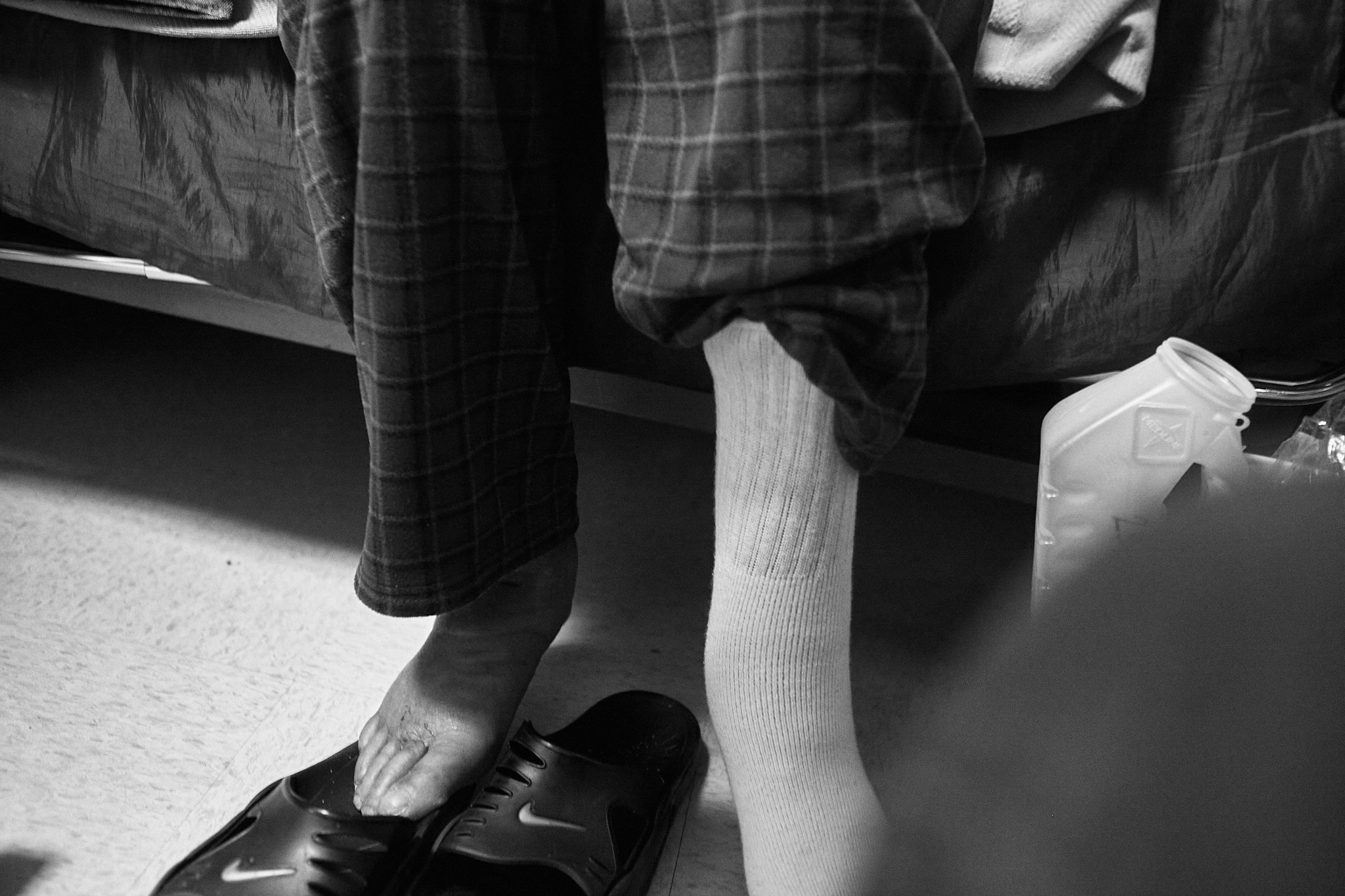 Diagonal view of a person sitting on a bed, with one foot bare and the other foot in a sock. The person is wearing loose plaid pants and a shirt, and a pair of Nike slide sandals are on the floor. A plastic jug and some plastic bags are on the floor 