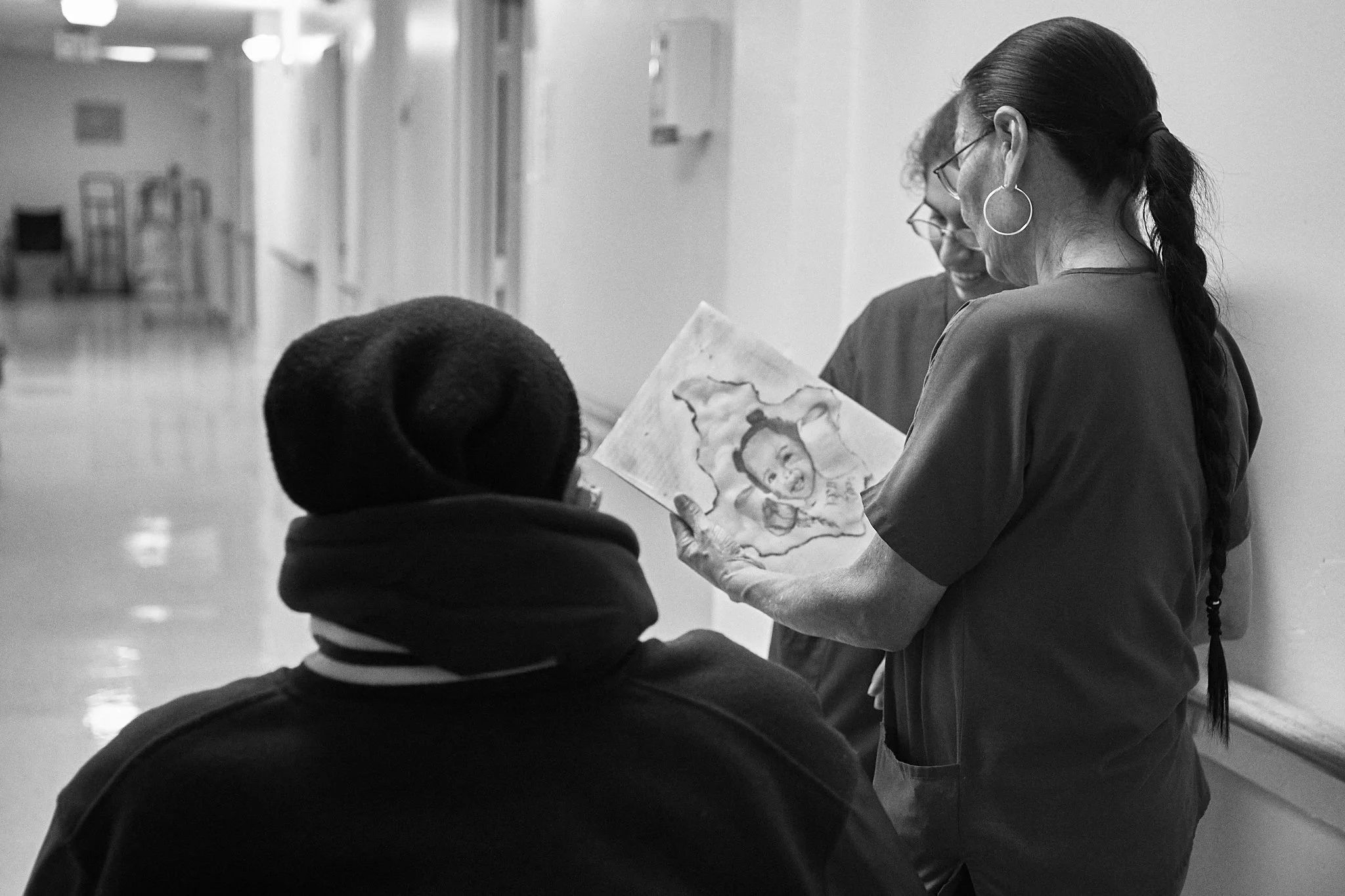 Two women and a child sitting in a hospital hallway, with the women looking at a picture of a smiling baby.