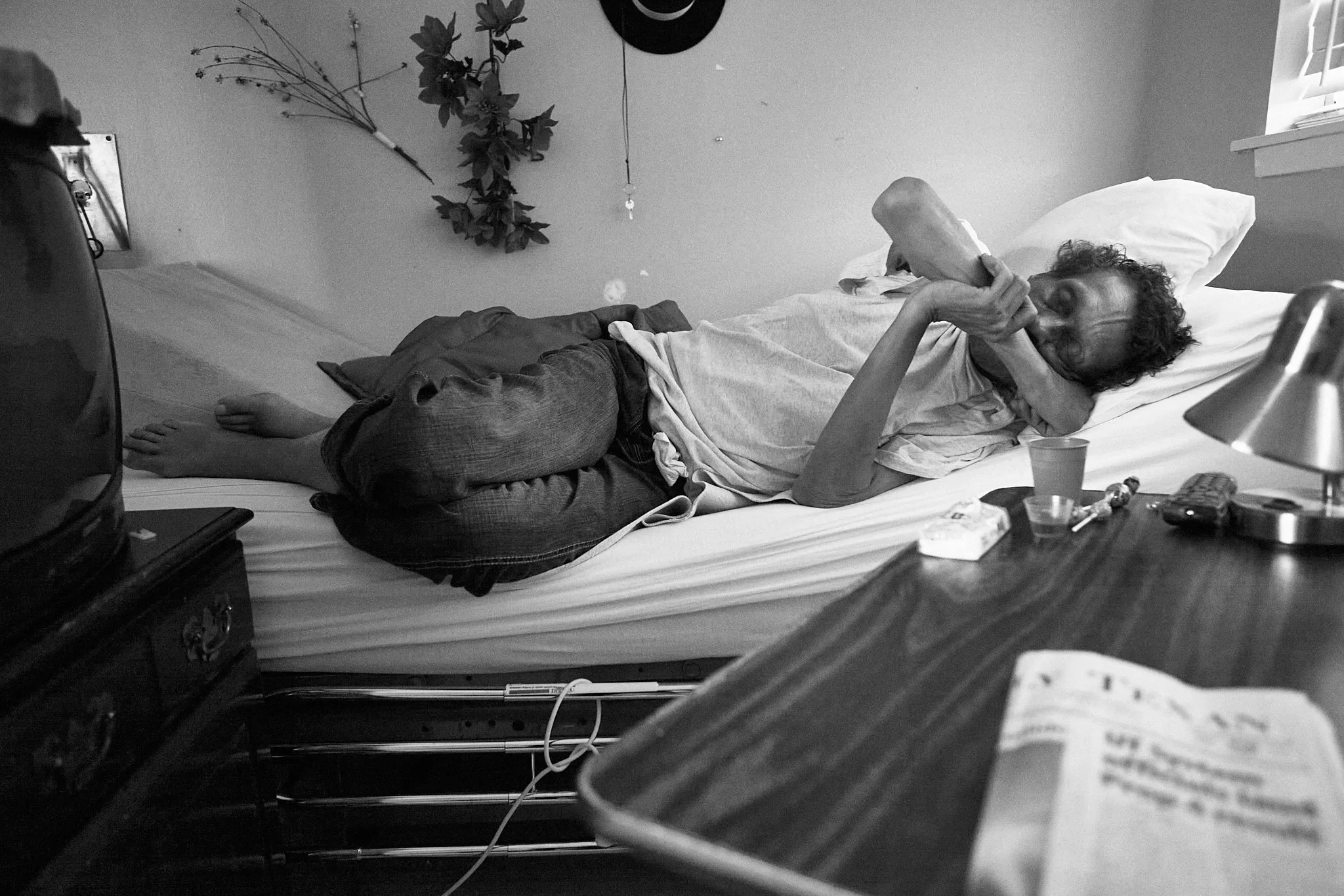 A person lying on a bed, drinking from a large bottle, in a bedroom with a plant, a hat hanging on the wall, and various items on a nightstand including a cup, medication, and a remote control.