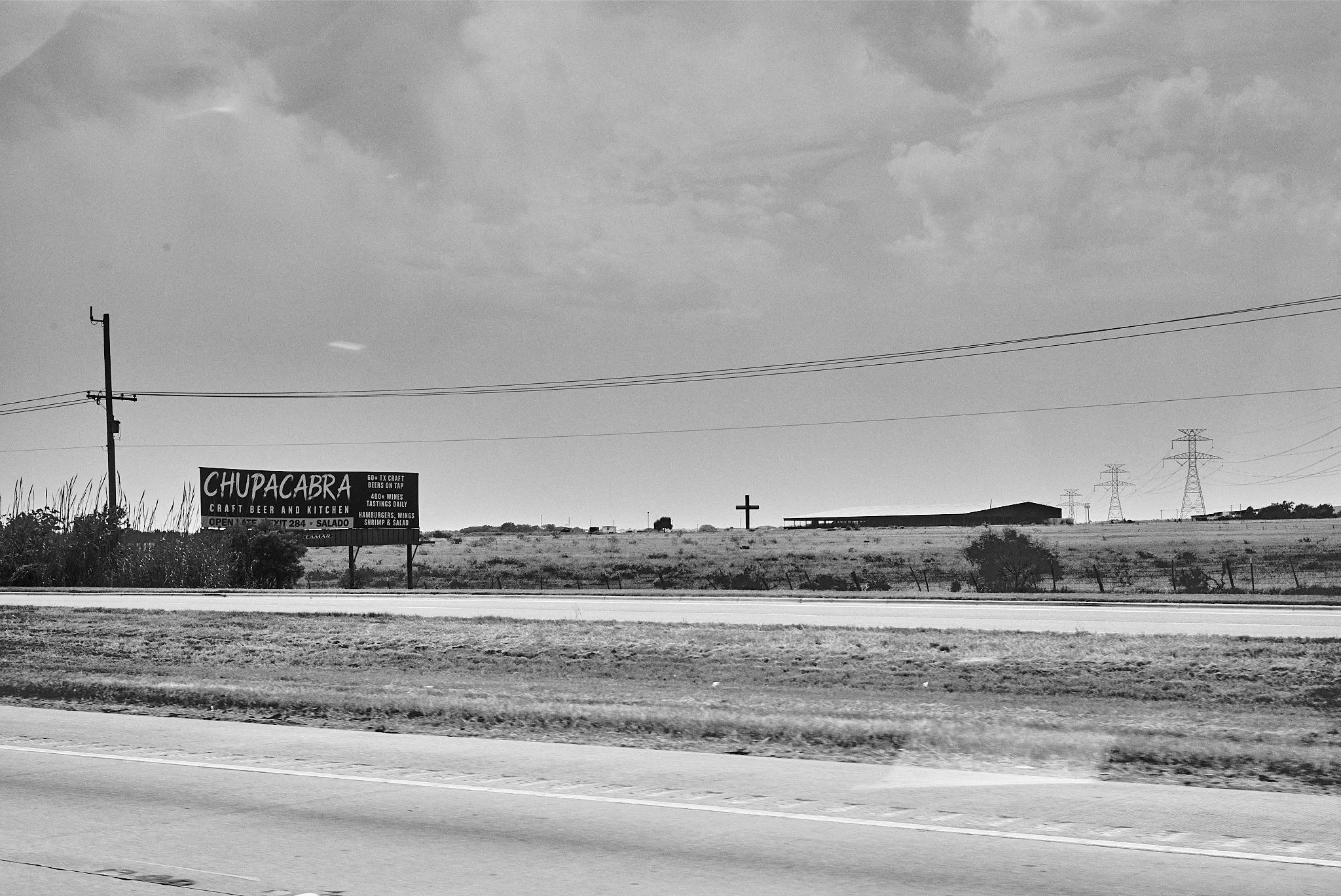 A black and white photo of a rural landscape viewed from a moving vehicle. There is a billboard advertising Chupacabra craft beer and kitchen, a cross on the hill, power lines, and a low building in the distance.