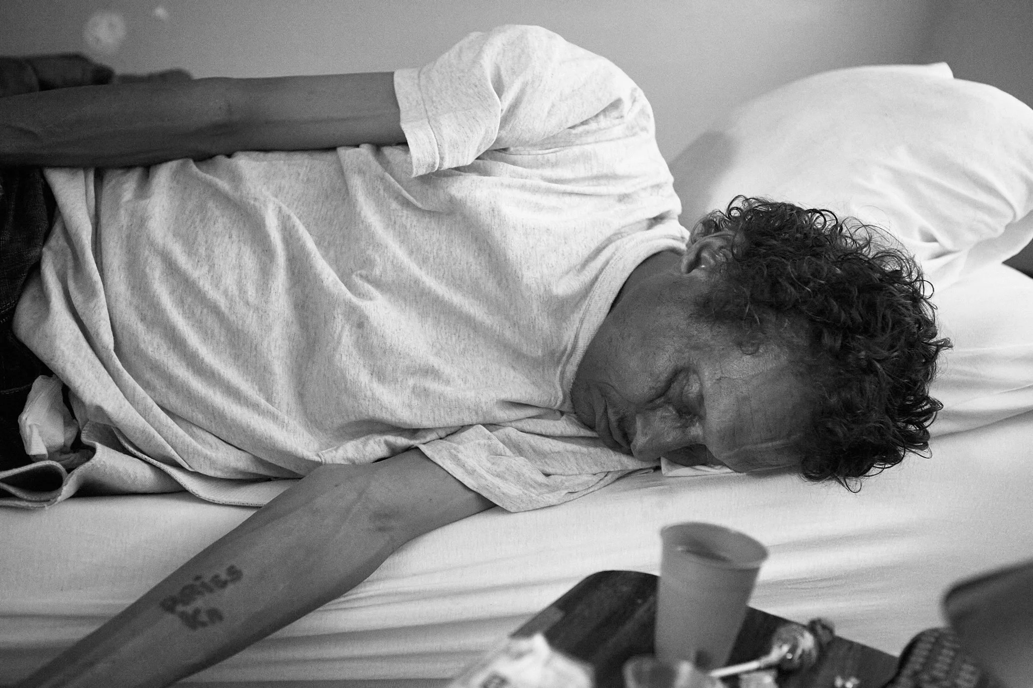 A man with curly hair sleeps on a bed with his arm stretched out and a tattoo visible on his forearm."