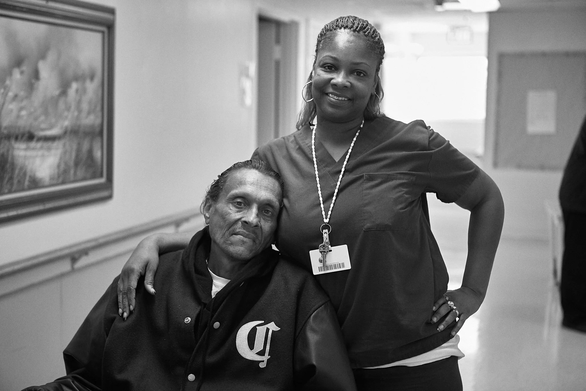 A woman dressed in medical scrubs with a nurse badge, standing next to a seated man wearing a black jacket with a letter 'C' on it. The woman has her arm around the man's shoulder and is smiling at the camera.