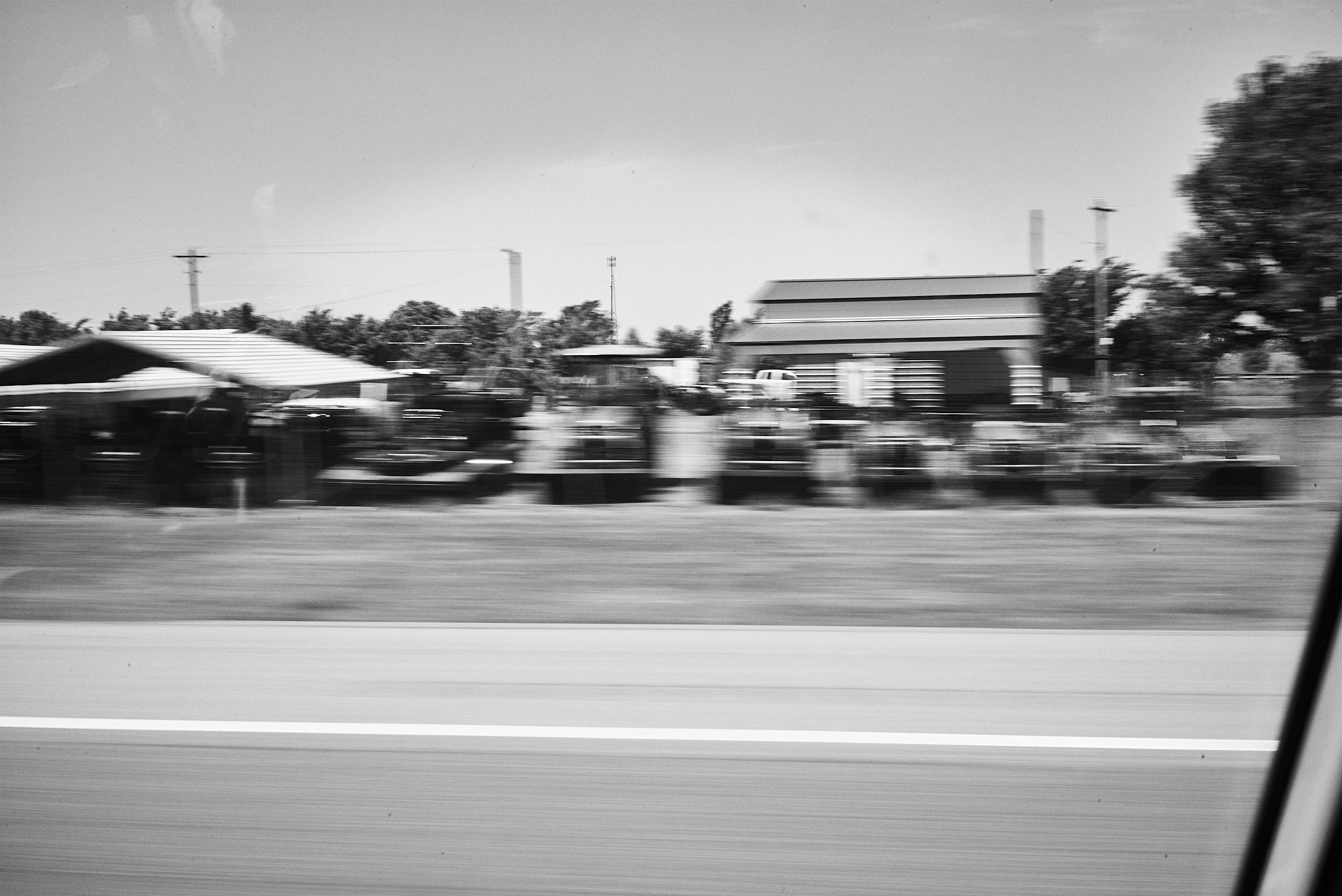 Blurry black-and-white photo of a rural area with houses, trees, and a horse barn seen from inside a moving vehicle.