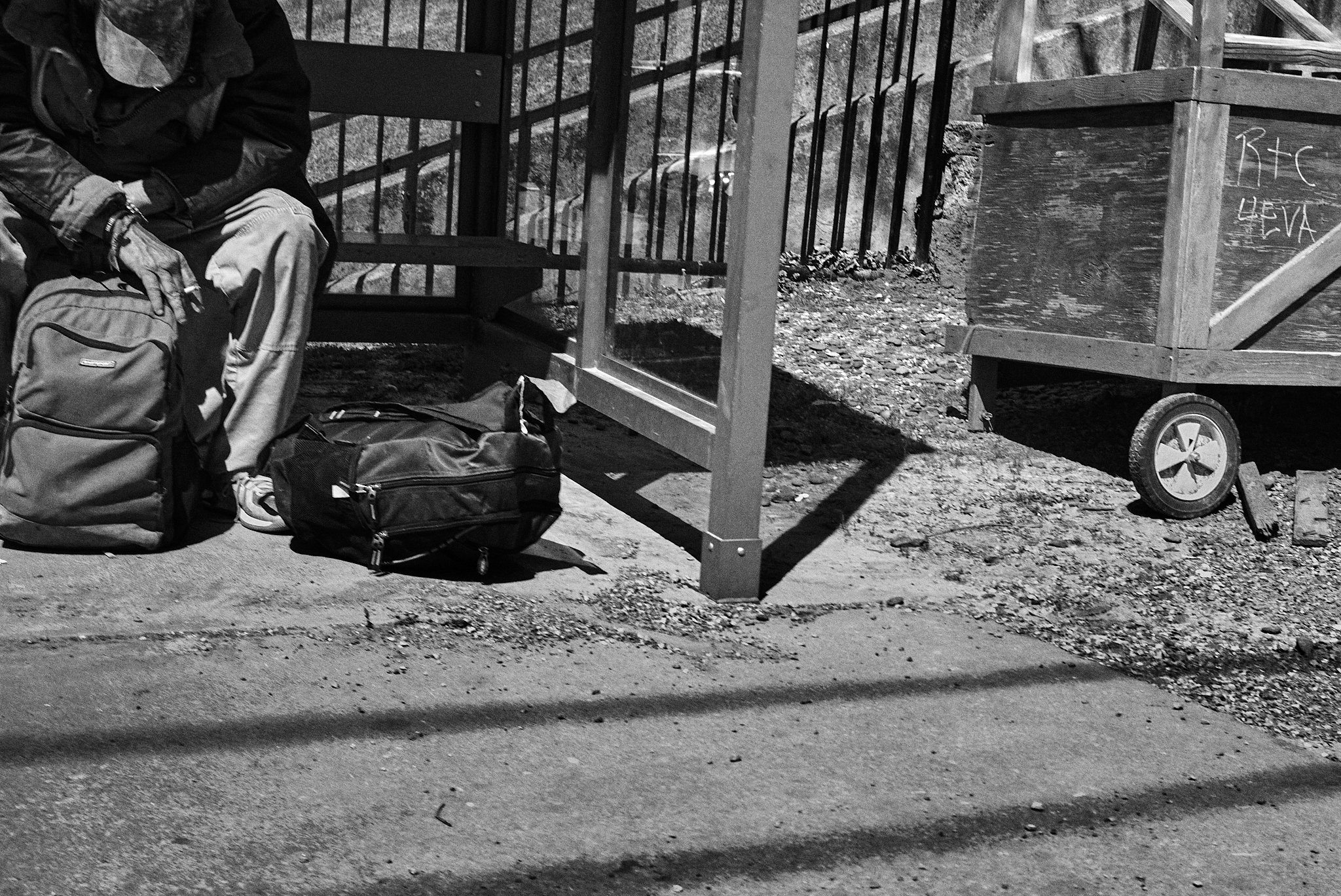 A person sitting on a bench with backpacks on the ground nearby, in front of a fenced and a wooden cart on an outdoor sidewalk.