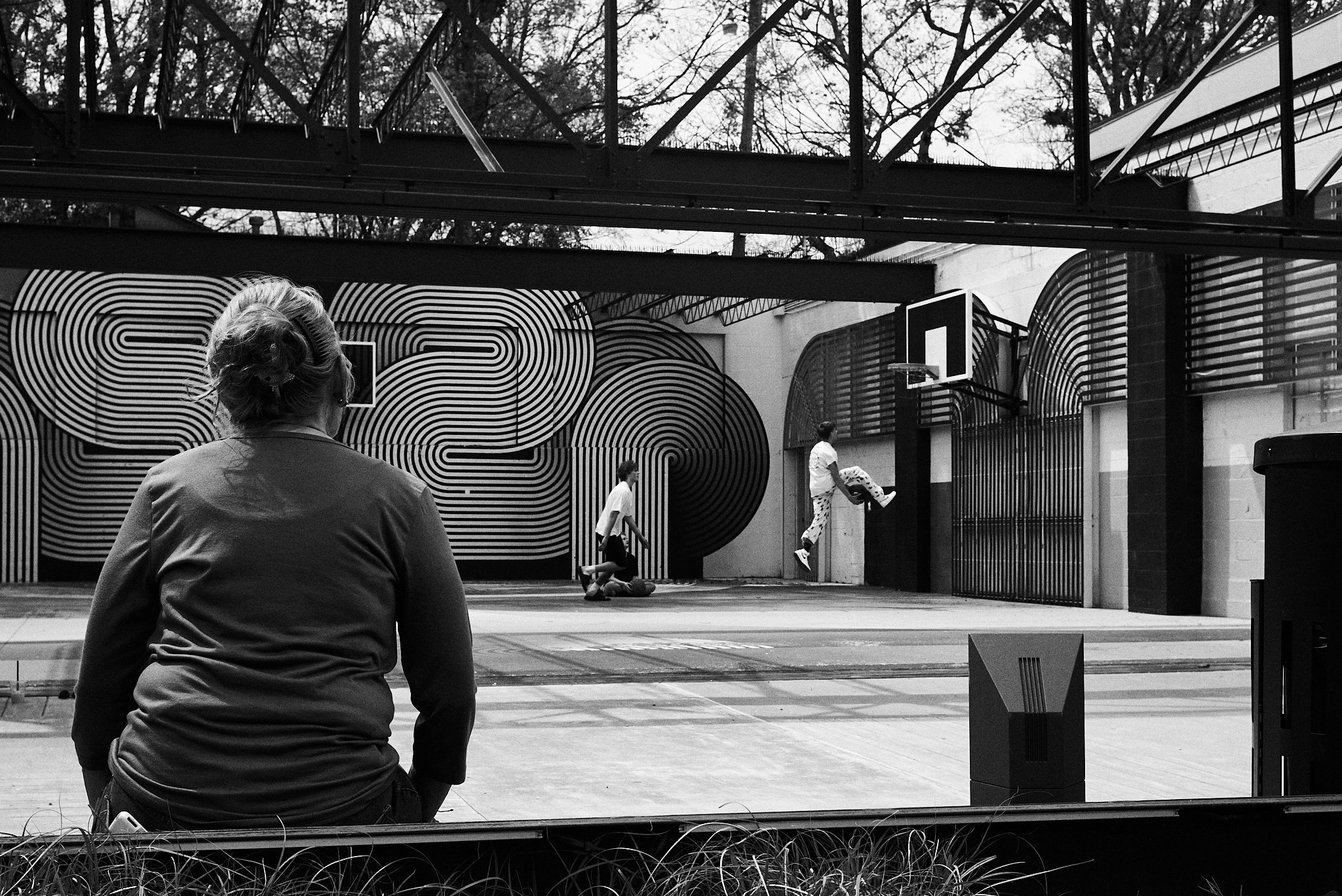 A woman sitting and watching two children playing basketball in an indoor half-court gym with a large, abstract black and white mural on the wall.