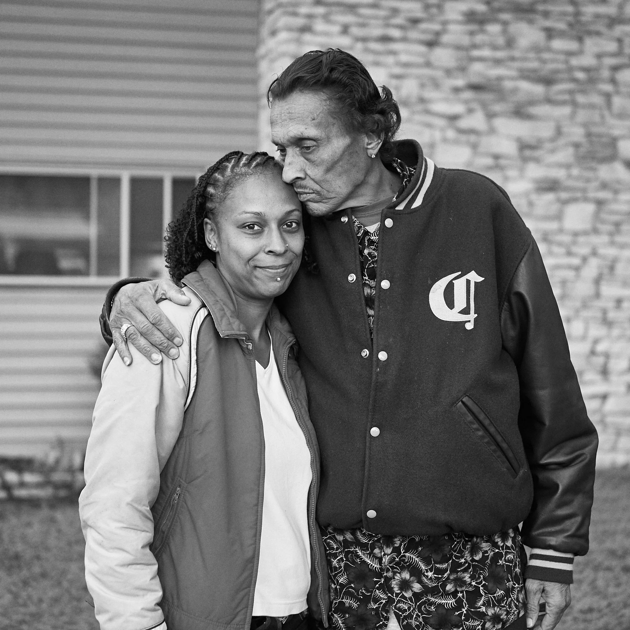 A black and white photo of two people, an older man and a younger woman, standing close together outdoors. The man is leaning his head on the woman's, and they are embracing. The woman has braids and is smiling slightly, while the man has his eyes cl