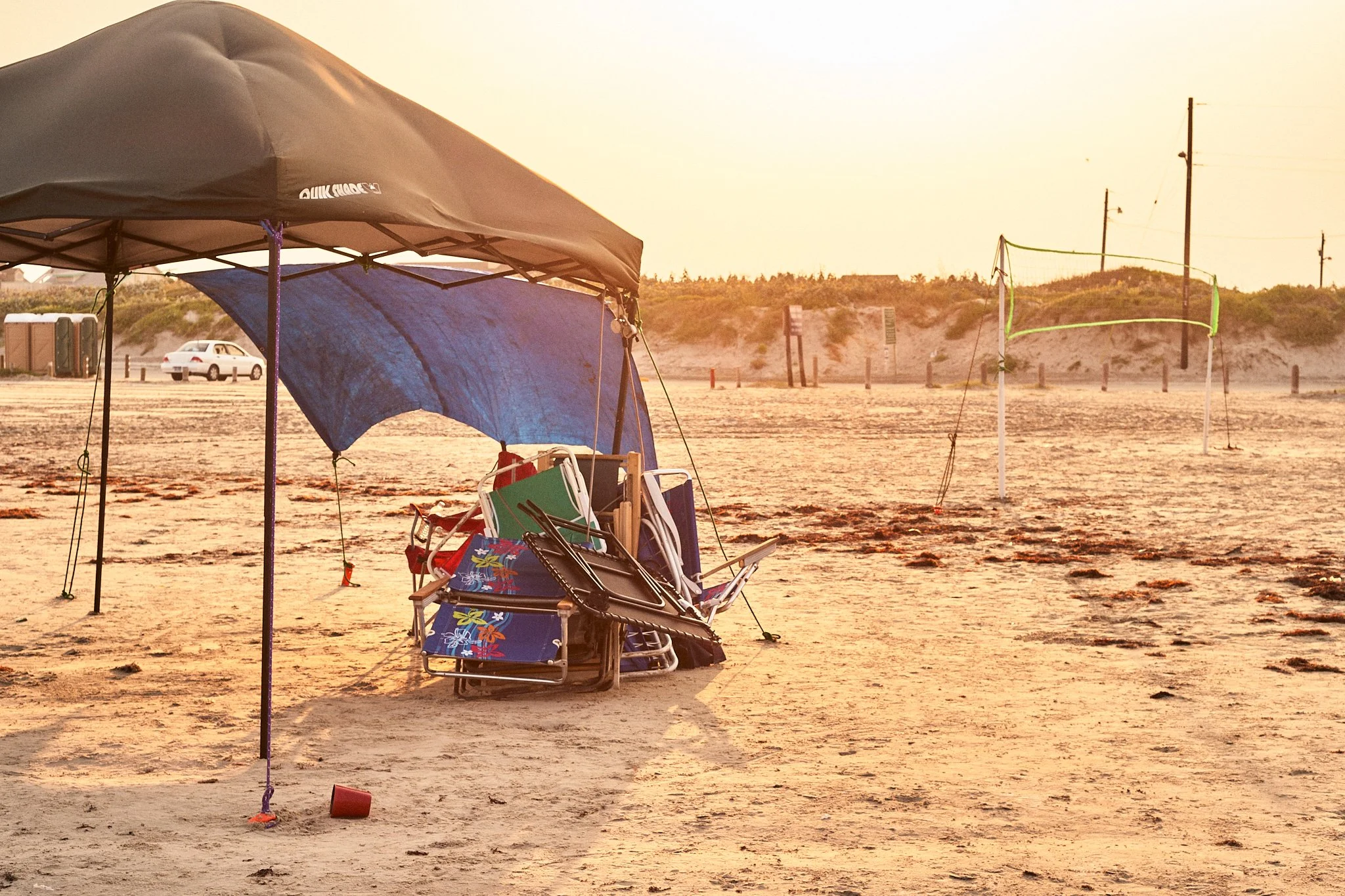 Empty beach with a large umbrella, beach chairs, and a blue towel near the shoreline. Volleyball net posts are visible in the distance, illuminated by warm sunset light.