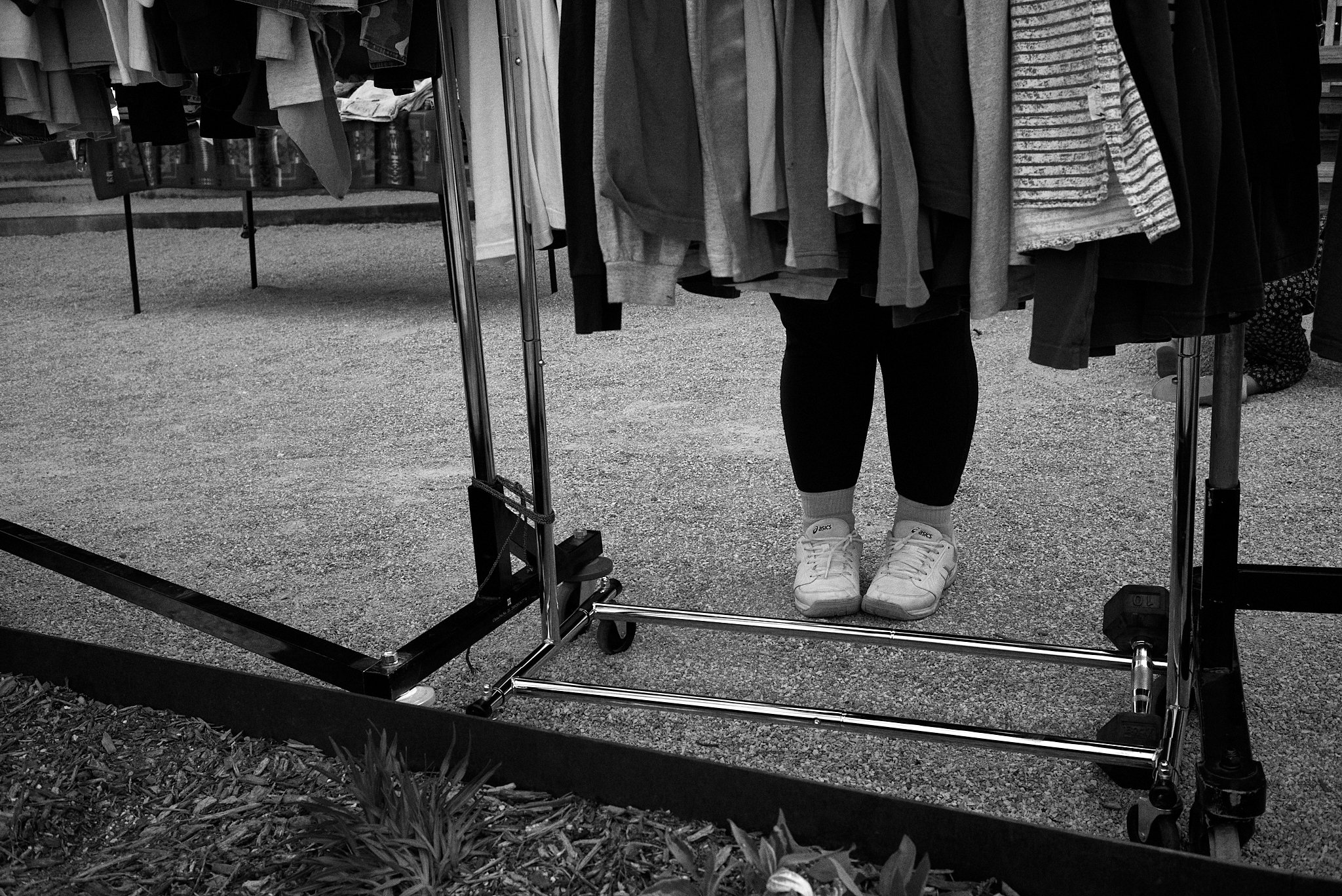 Person standing behind a clothing rack, with only their legs visible, wearing sneakers and black leggings, at an outdoor market.