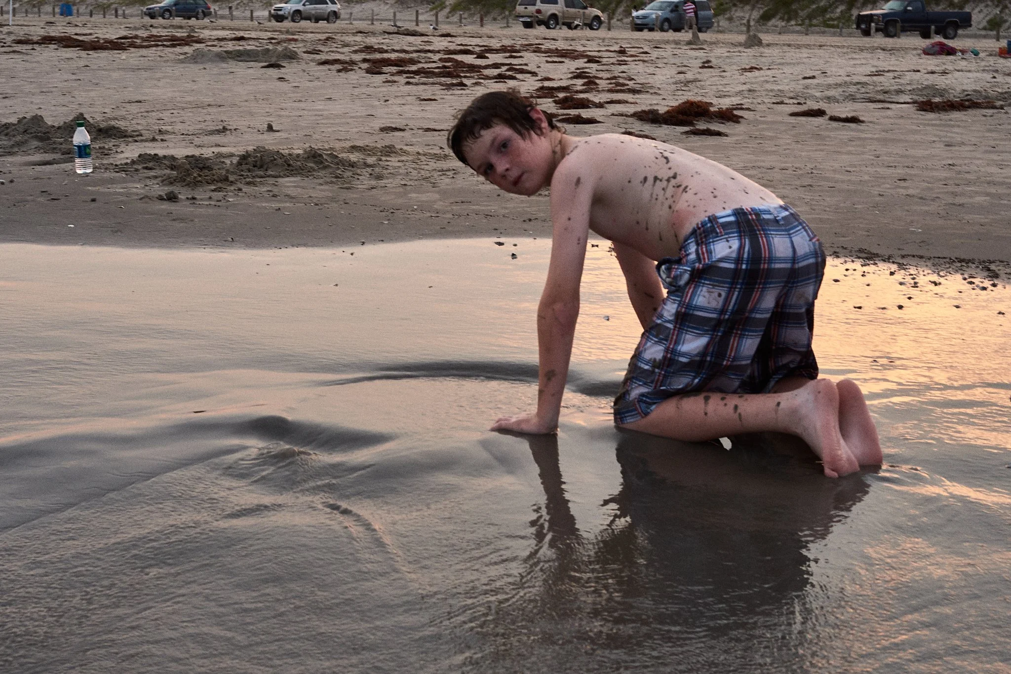 A young boy kneeling on the sand near the shoreline at a beach, with his hands in the water and looking at the camera, wearing plaid shorts and no shirt, with footprints and boats in the background.