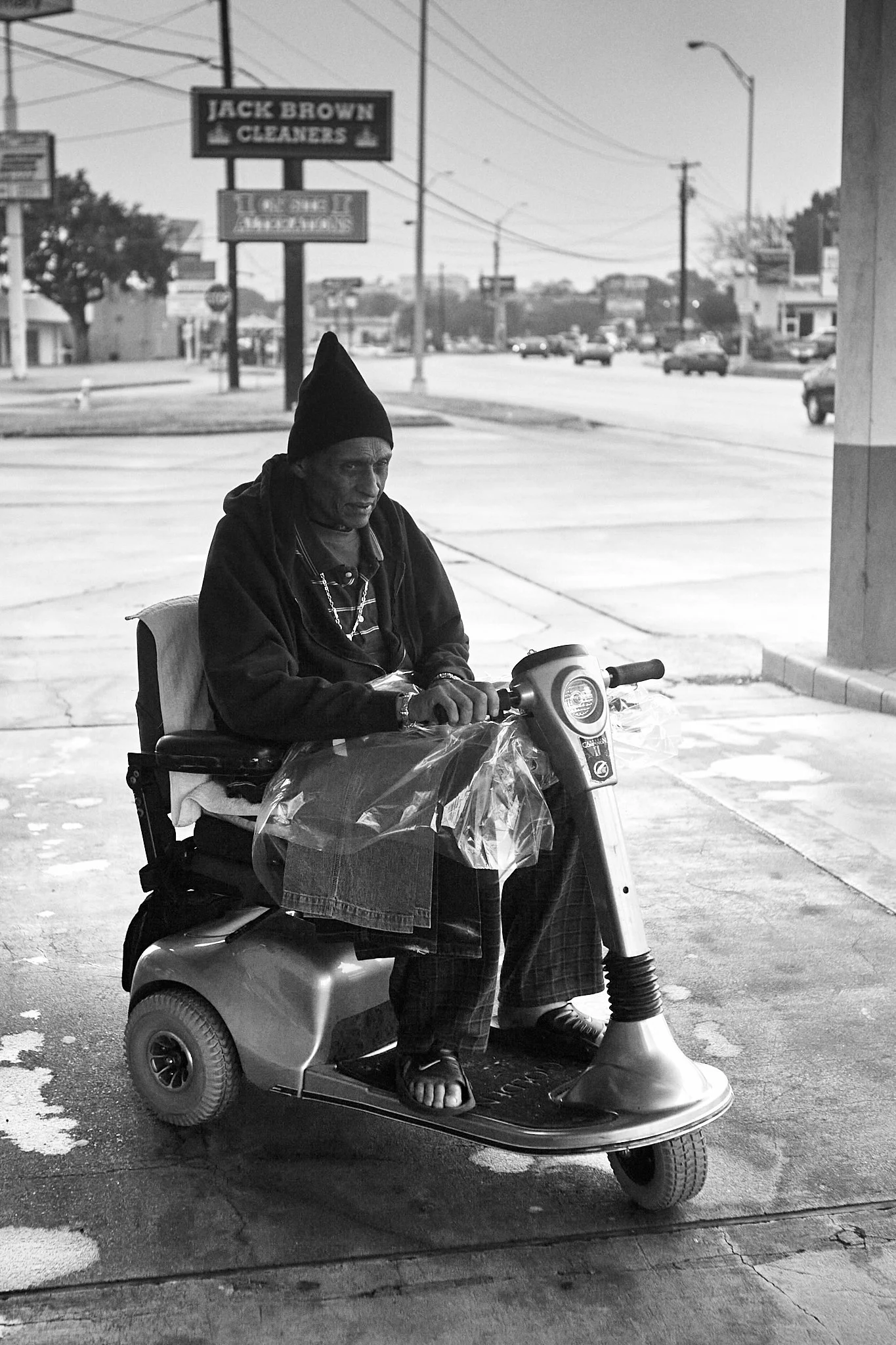 An elderly man wearing a beanie hat, sitting on a mobility scooter, is parked outdoors on a sidewalk. He is dressed in dark clothing, with a jacket and jeans. A plastic bag and a piece of cloth are attached to the scooter. In the background, there is
