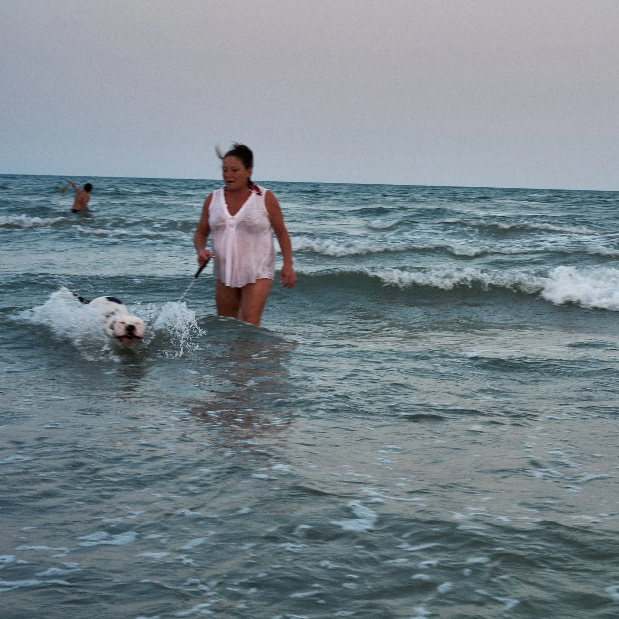 A woman walking in the ocean waves with a dog on a leash, another person swimming in the background, at the beach during the evening.