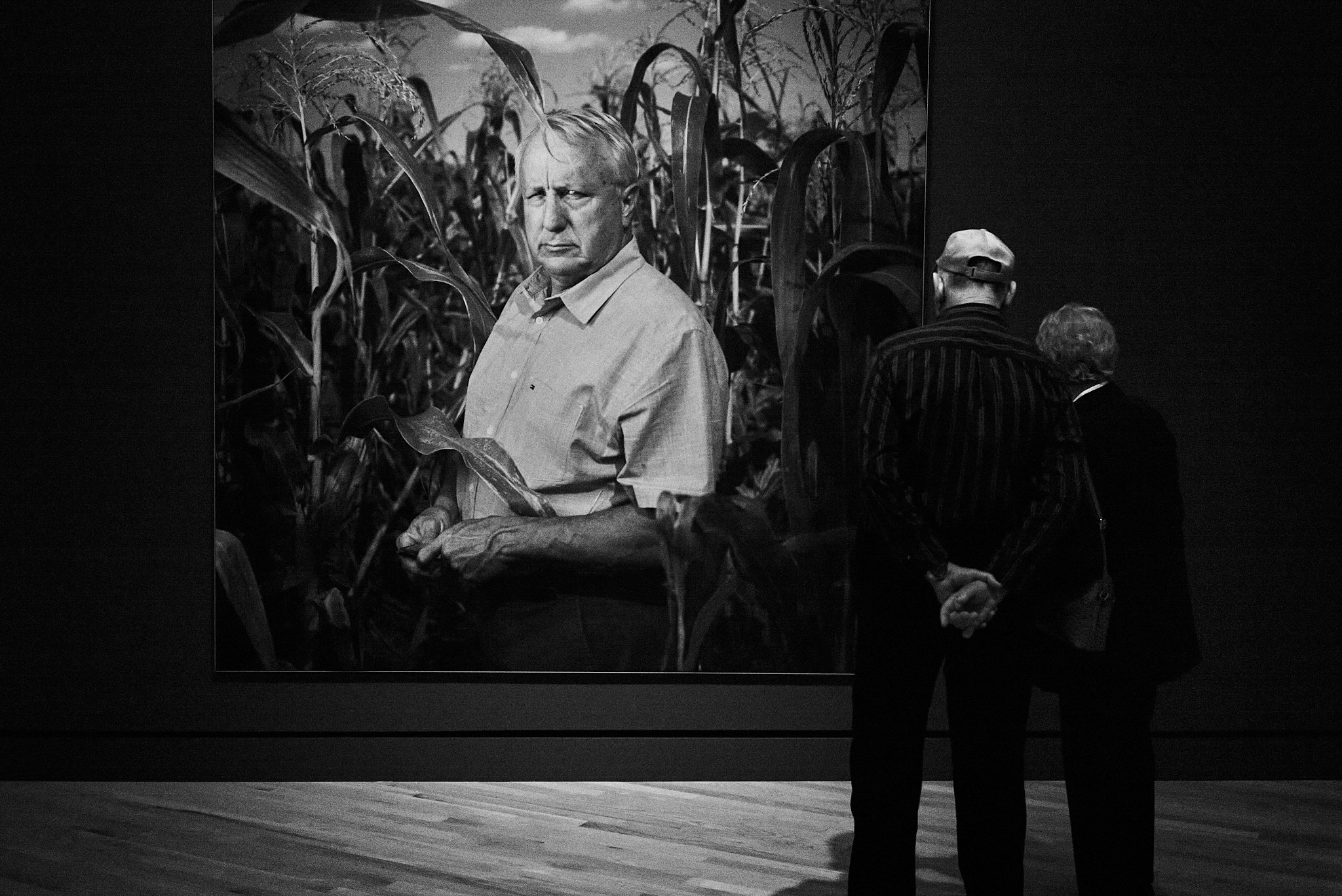 Two people standing in an art gallery, observing a black and white photograph of a man in a field of tall plants, possibly corn.