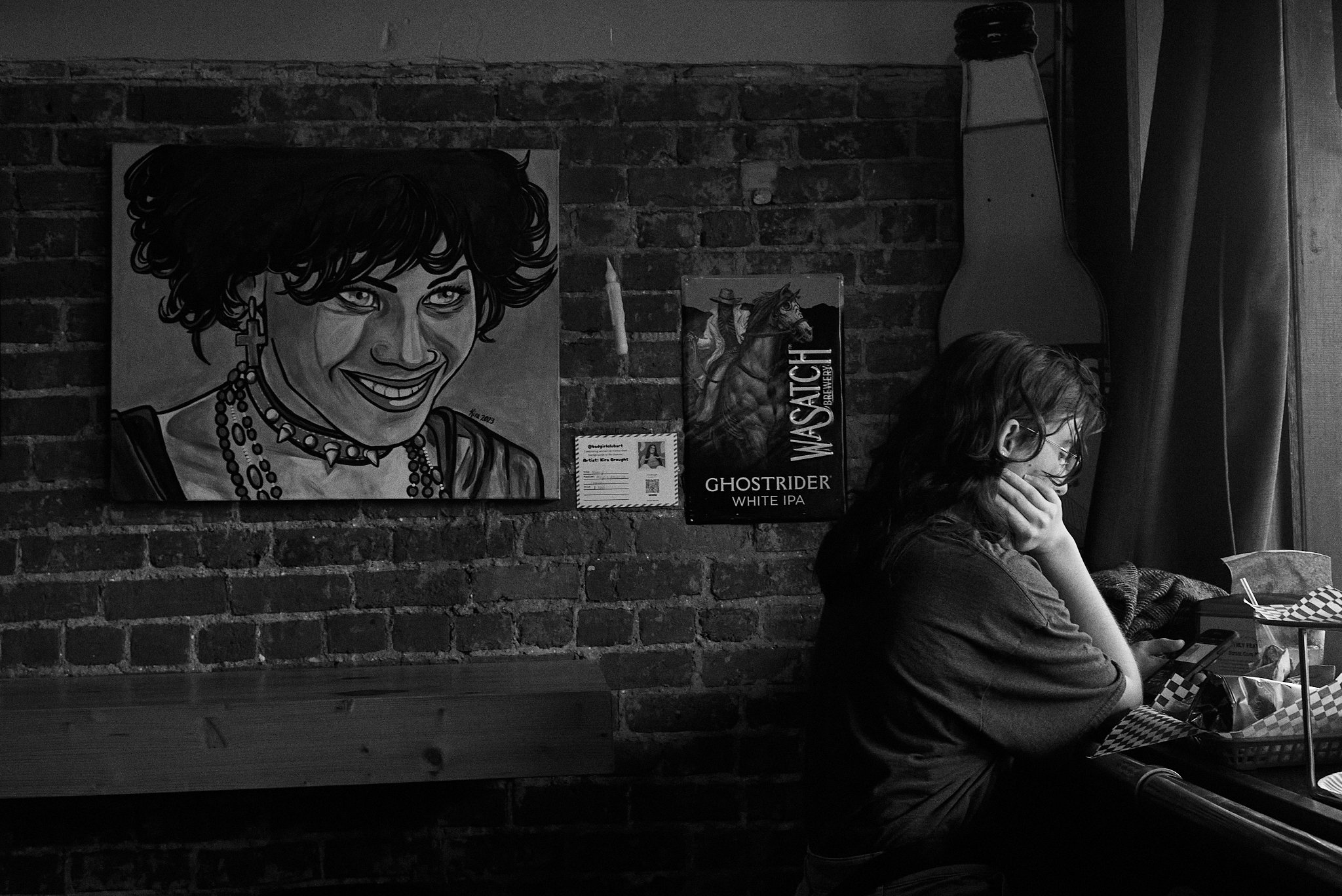 A woman sitting at a table, resting her chin on her hand, with her phone in front of her, in a room with brick walls decorated with posters, including one of a smiling woman with earrings and flowers in her hair.