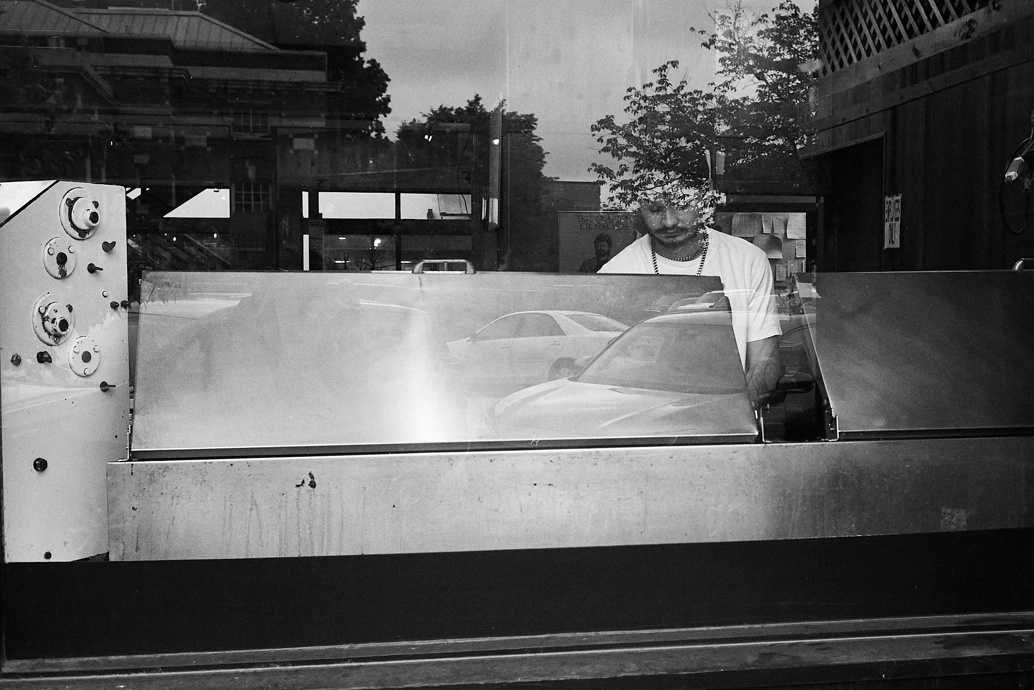 A person wearing a white T-shirt and a gold chain is working behind a glass display case, reflecting cars and trees outside on a city street.