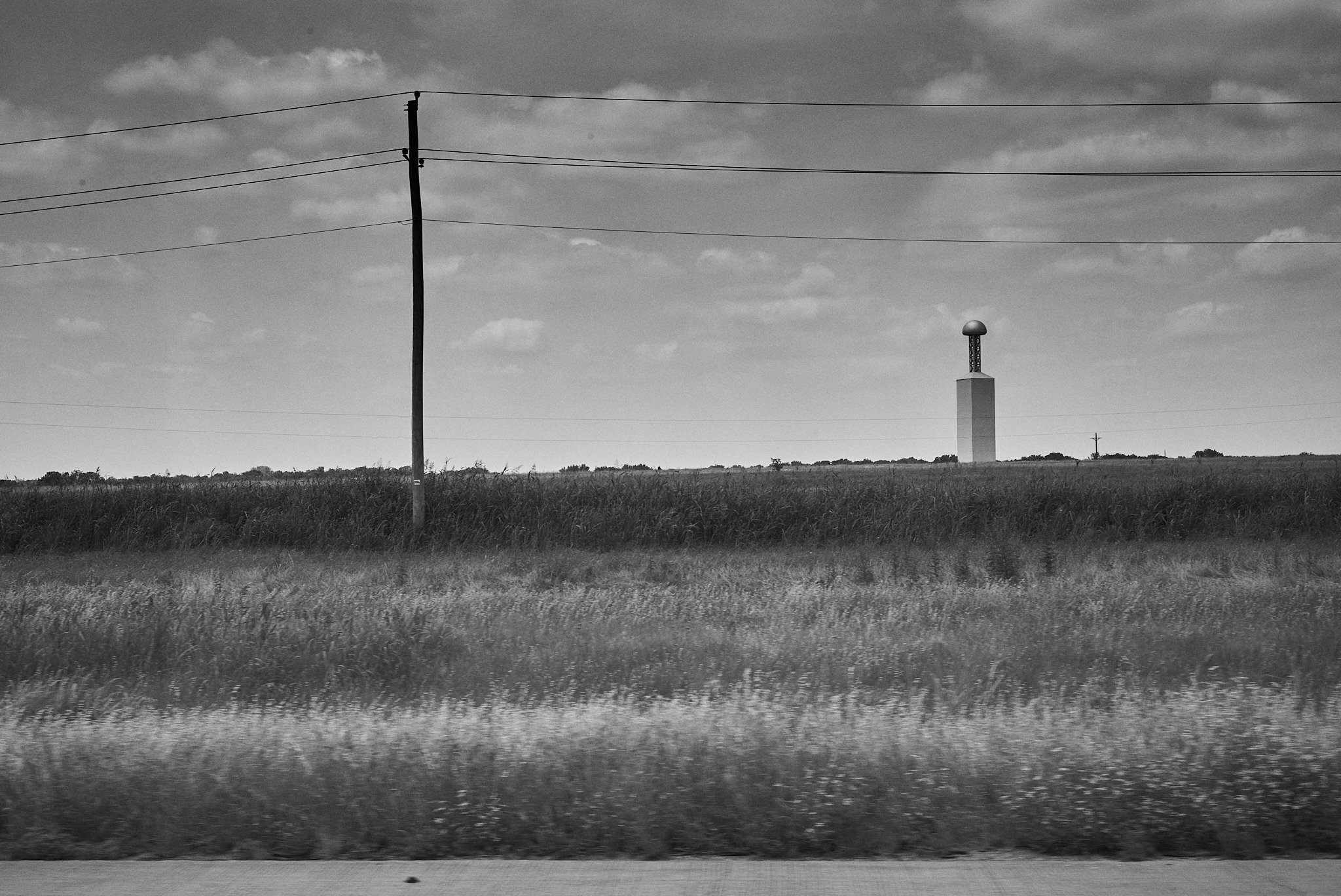Black and white photo of an open field with tall grass, a power pole with wires, and a tower with a spherical structure on top in the distance under a partly cloudy sky.