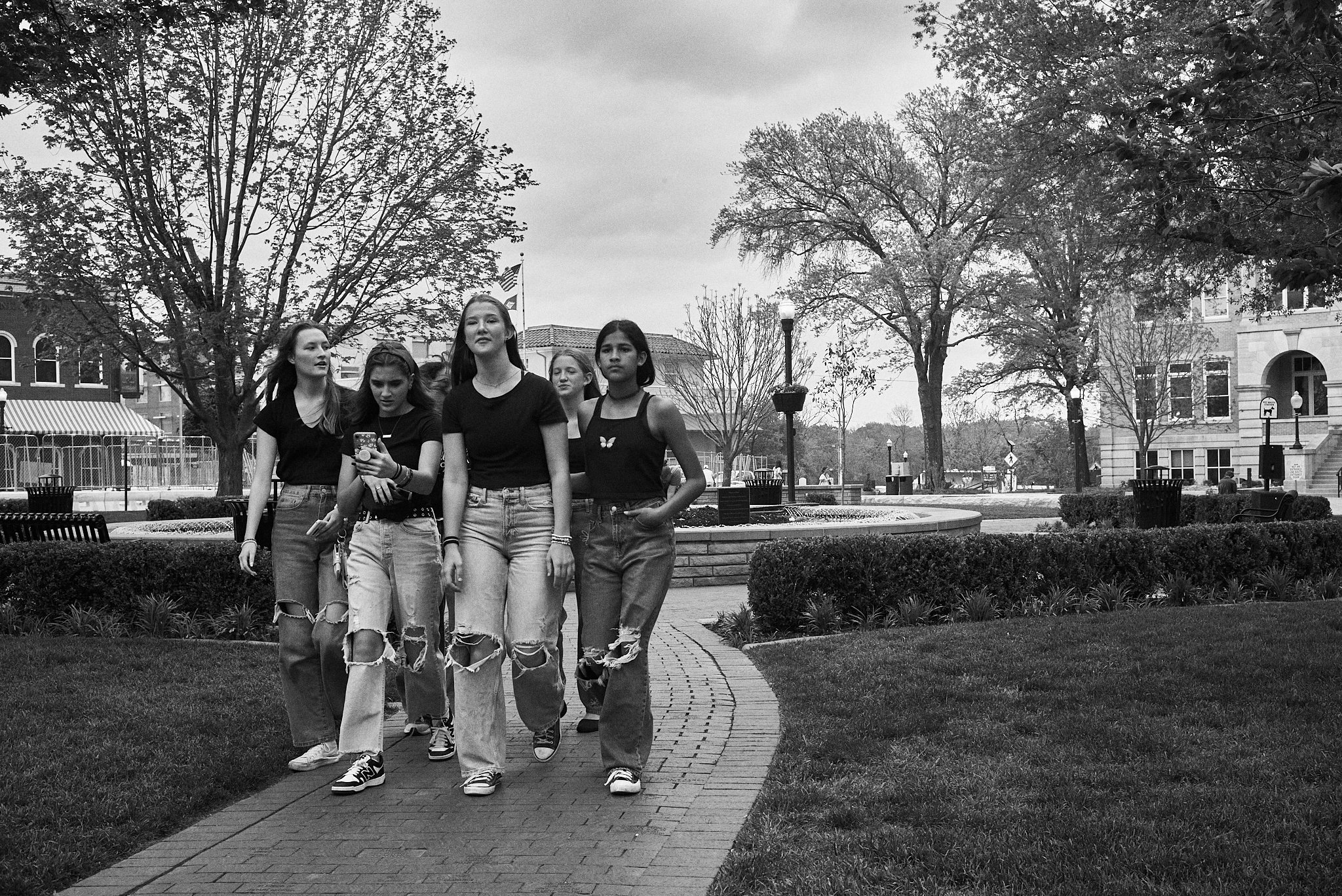 Five young women walking on a paved path in a park, with trees, benches, and buildings in the background on a cloudy day.