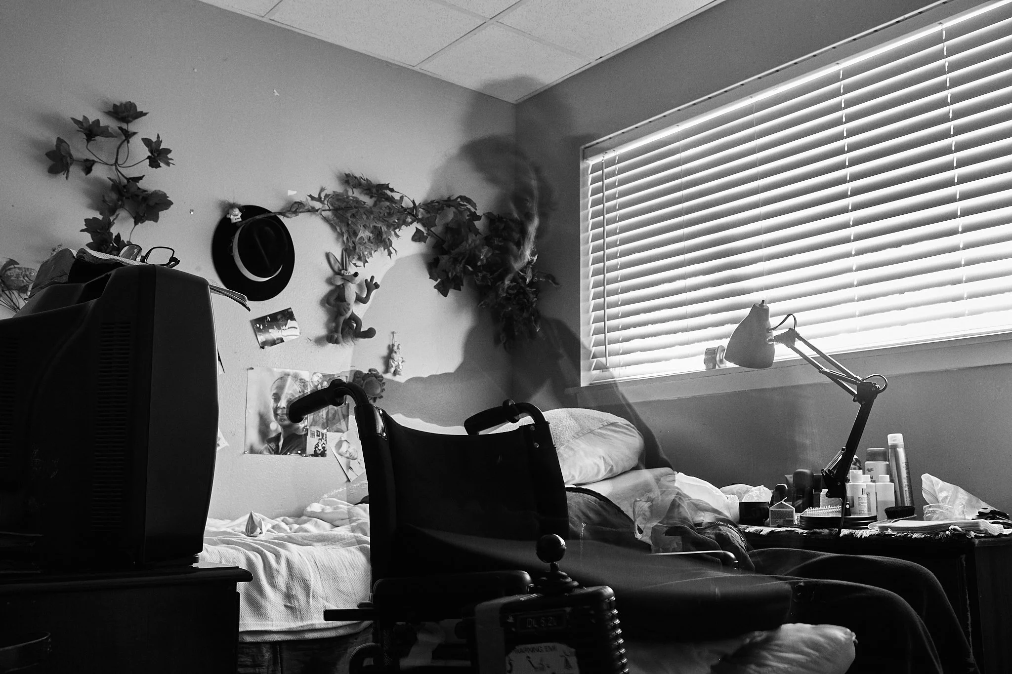 A black and white photo of a bedroom with sunlight coming through window blinds. A person is lying on the bed, with a ghostly, transparent image of their head and upper body visible, creating a dual-exposure effect.