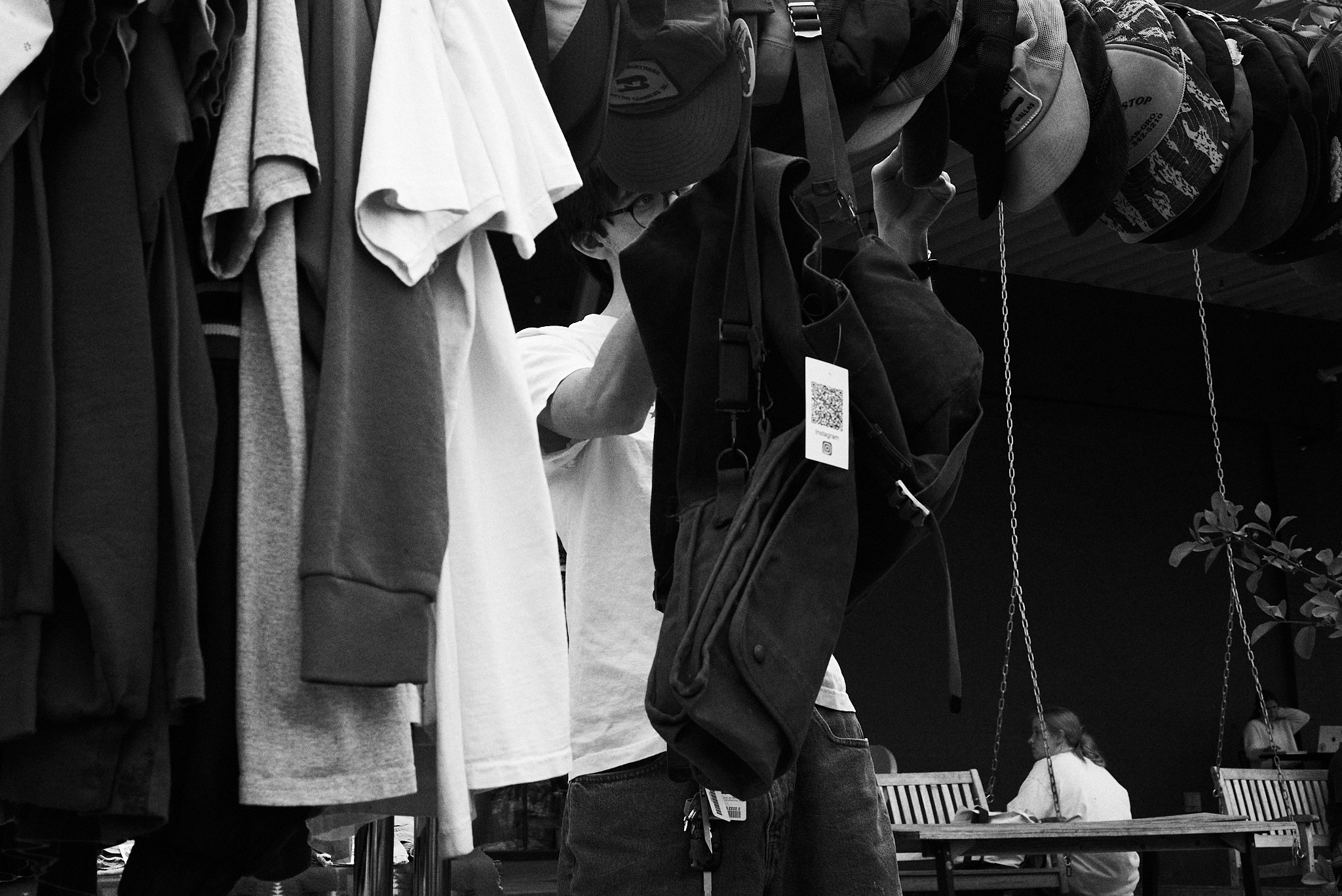 A person shopping for clothing and accessories at an indoor store, with a focus on hanging hats and backpacks, as other shoppers are seated in the background.