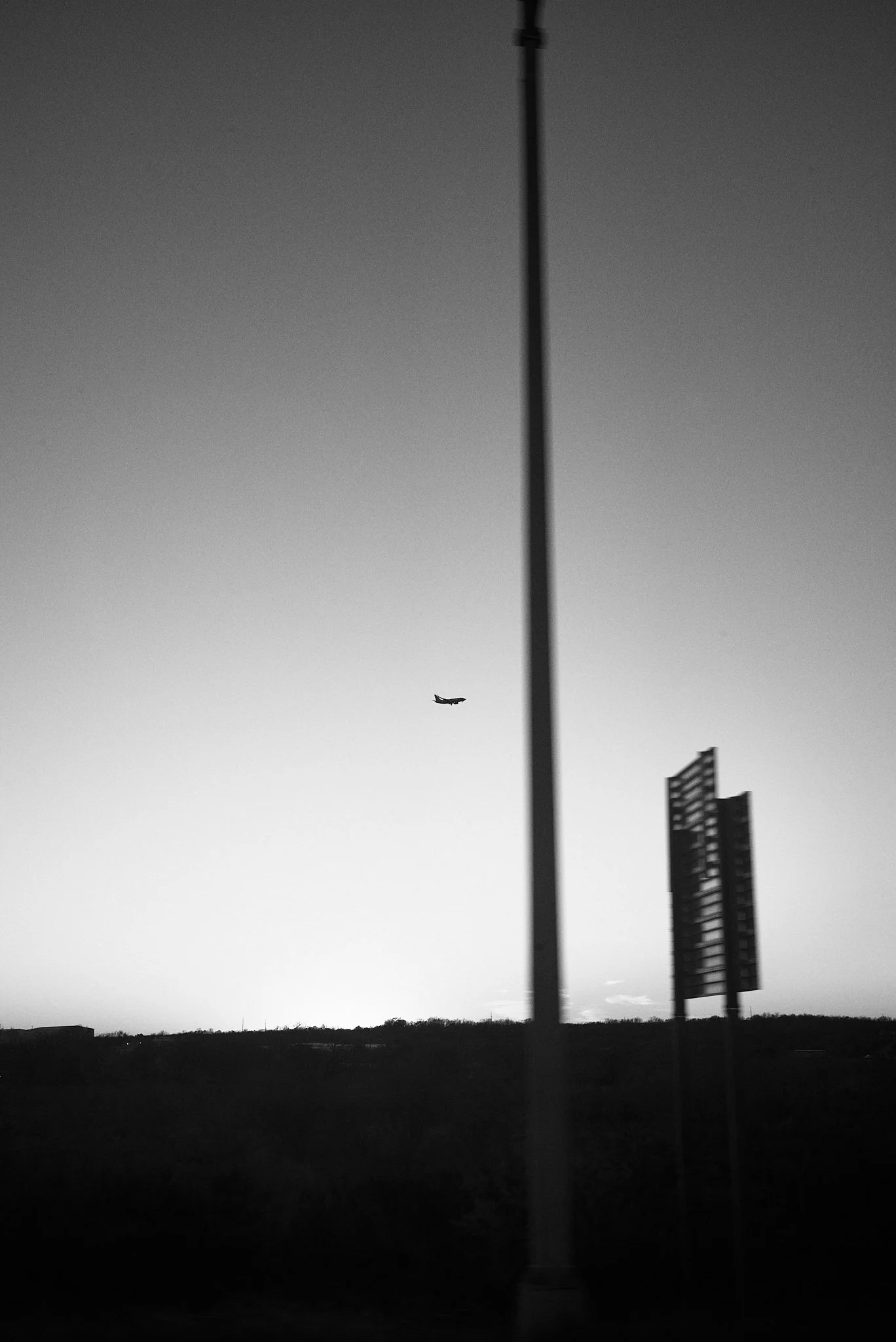 Black and white photo of a lone airplane flying in the sky above a landscape with a tall streetlight pole in the foreground and a building structure on the right side.