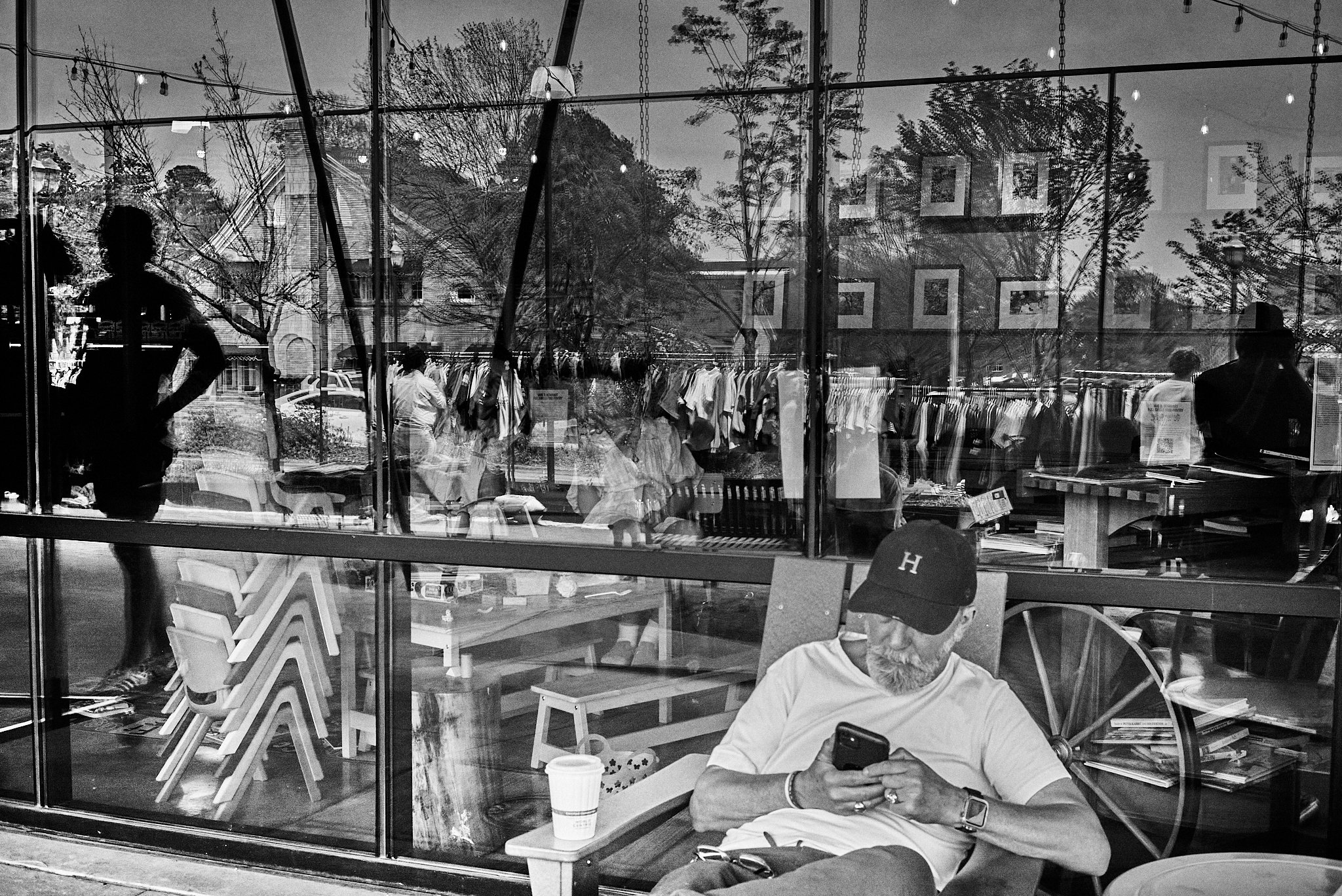 A black and white photo of a man with a beard and glasses sitting outside a storefront window, looking at his phone with a coffee cup on a nearby table. The window reflects trees, cars, and pedestrians walking by.