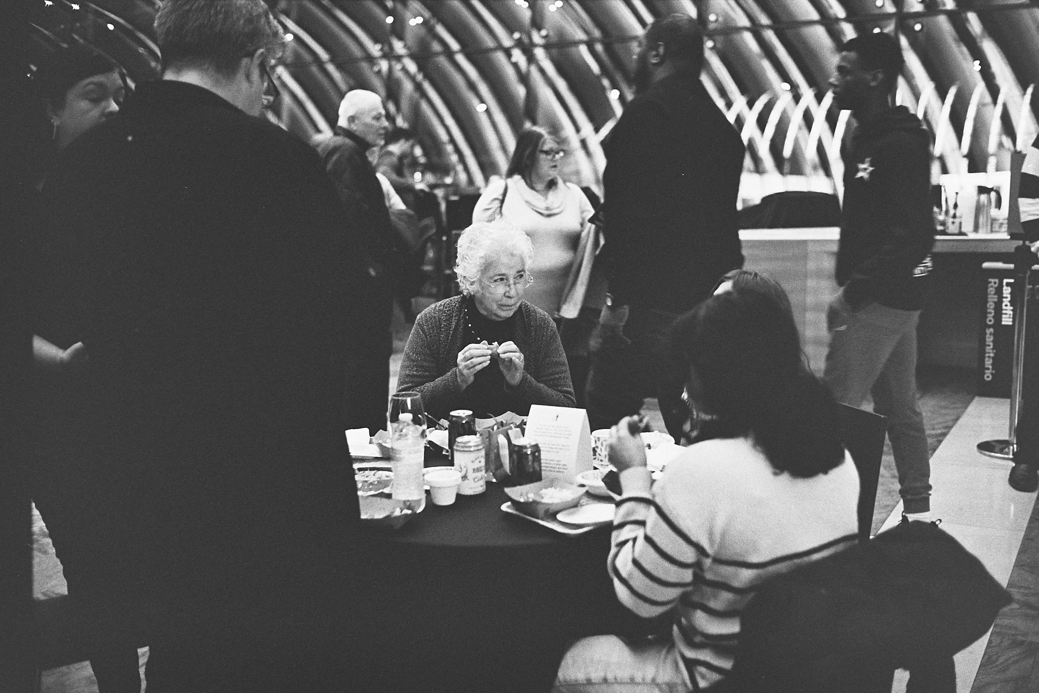 A group of people gathered around a table at an indoor setting, possibly a restaurant or airport lounge, with some standing and others seated, including an elderly woman with white curly hair sitting at the table and a woman with dark hair wearing a 