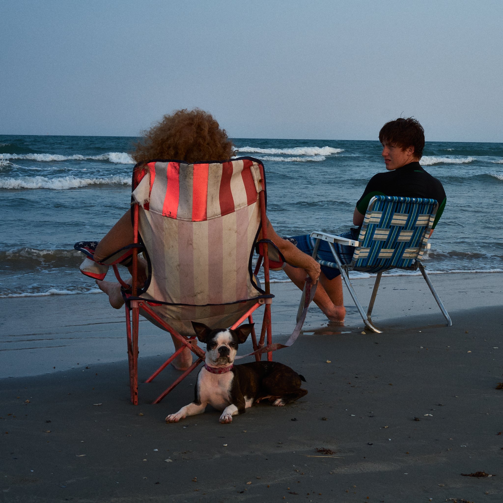 Two people sit on beach chairs near the ocean with a dog lying on the sand in front of them, during sunset or early evening.