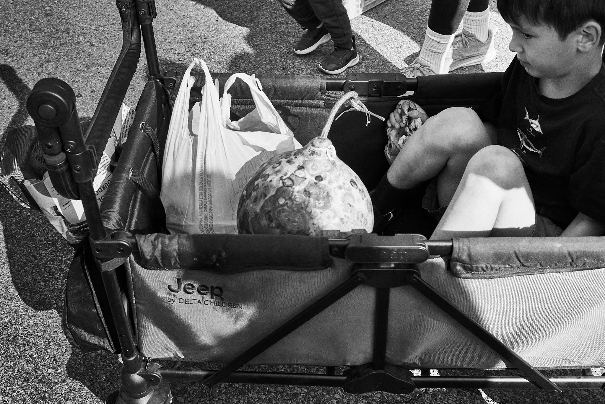 A young boy sitting in a wagon with a large gourd and a plastic shopping bag inside. Several children are visible, one standing nearby, all outdoors on a paved surface.