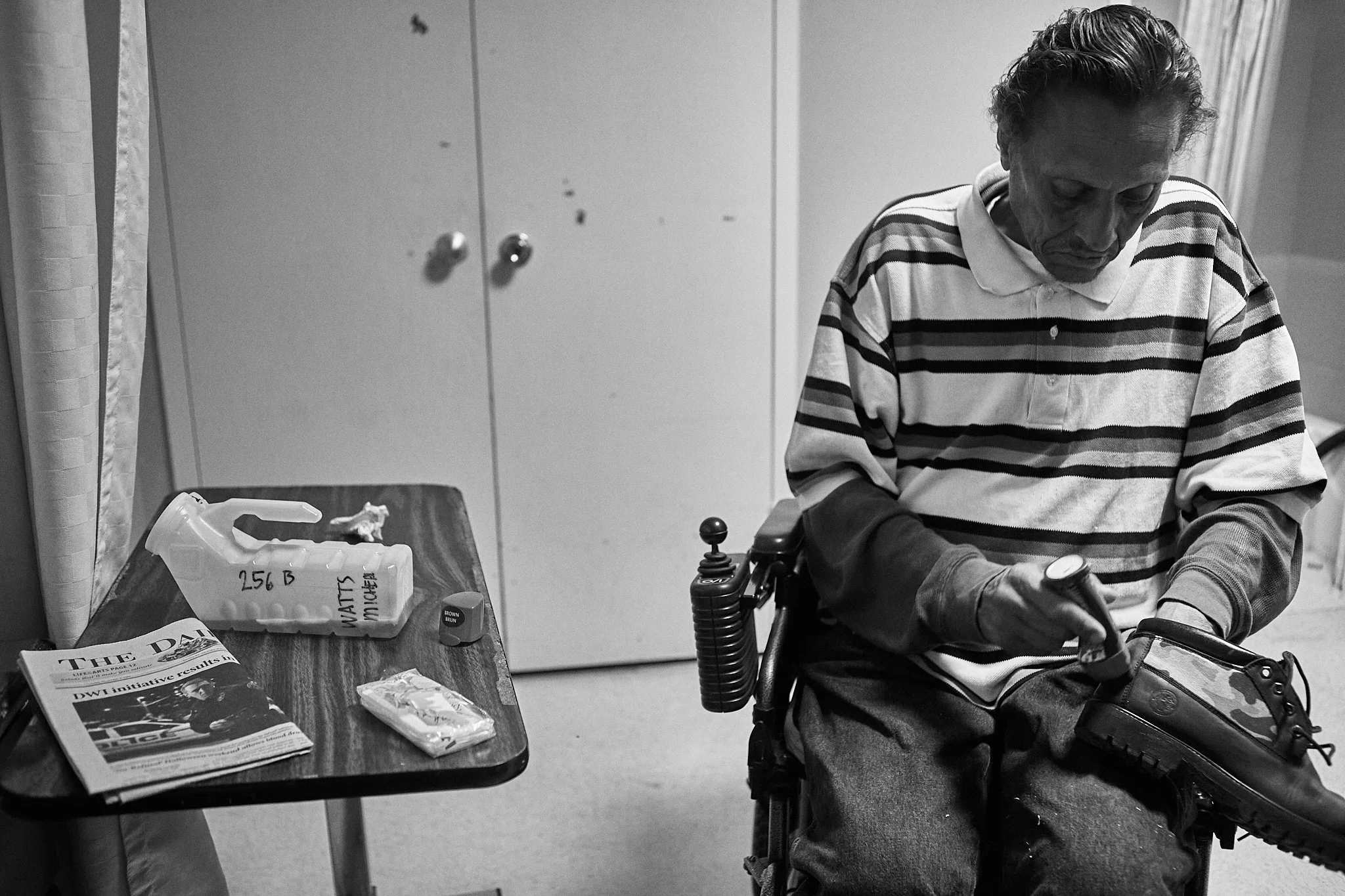 A man in a wheelchair holding a shoe, with a table beside him containing a newspaper, a small box, and tissues, in an indoor setting.