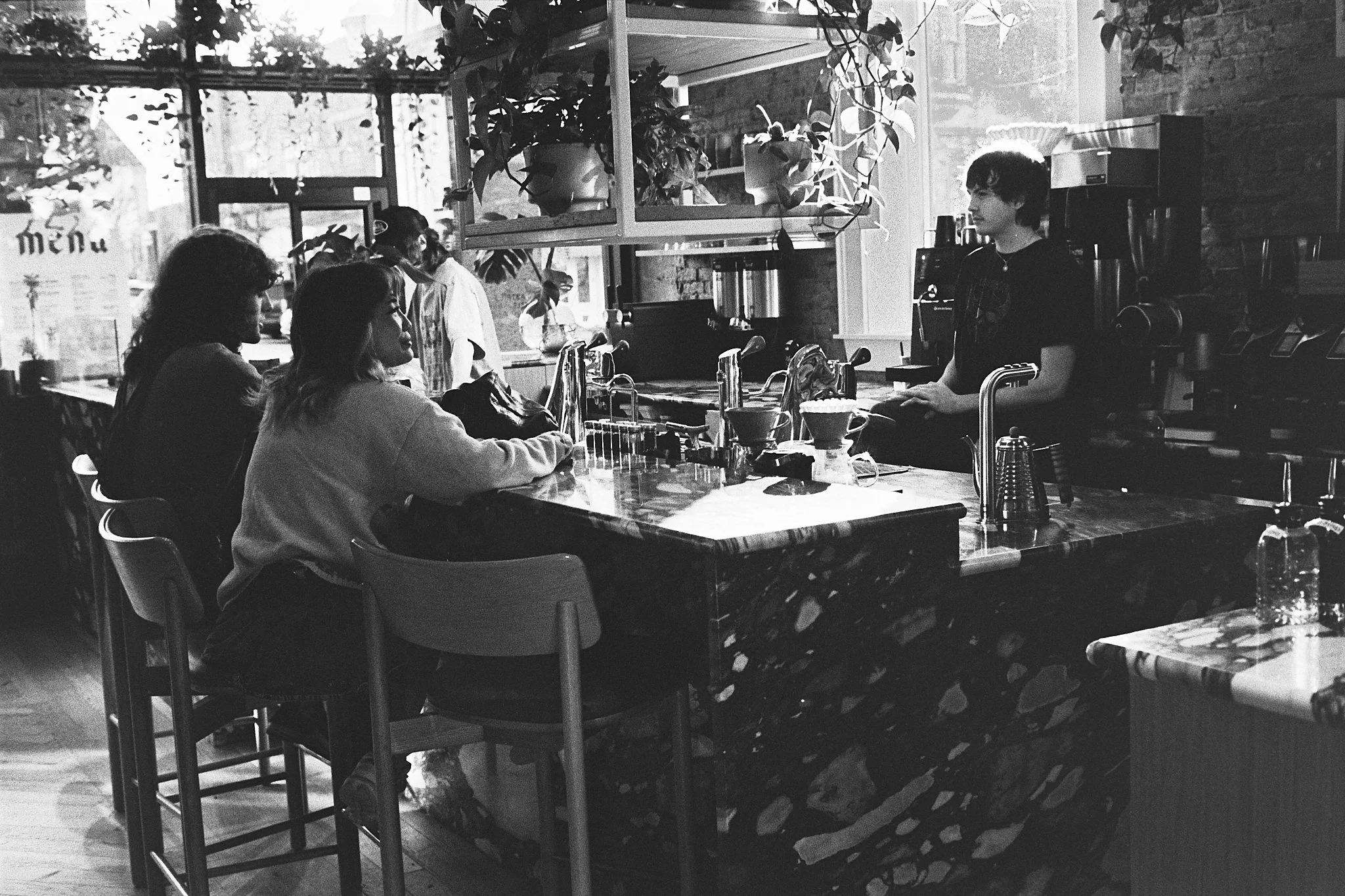 A black-and-white photo of a coffee shop interior with a barista behind the counter and three customers sitting on bar stools, engaging in conversation.