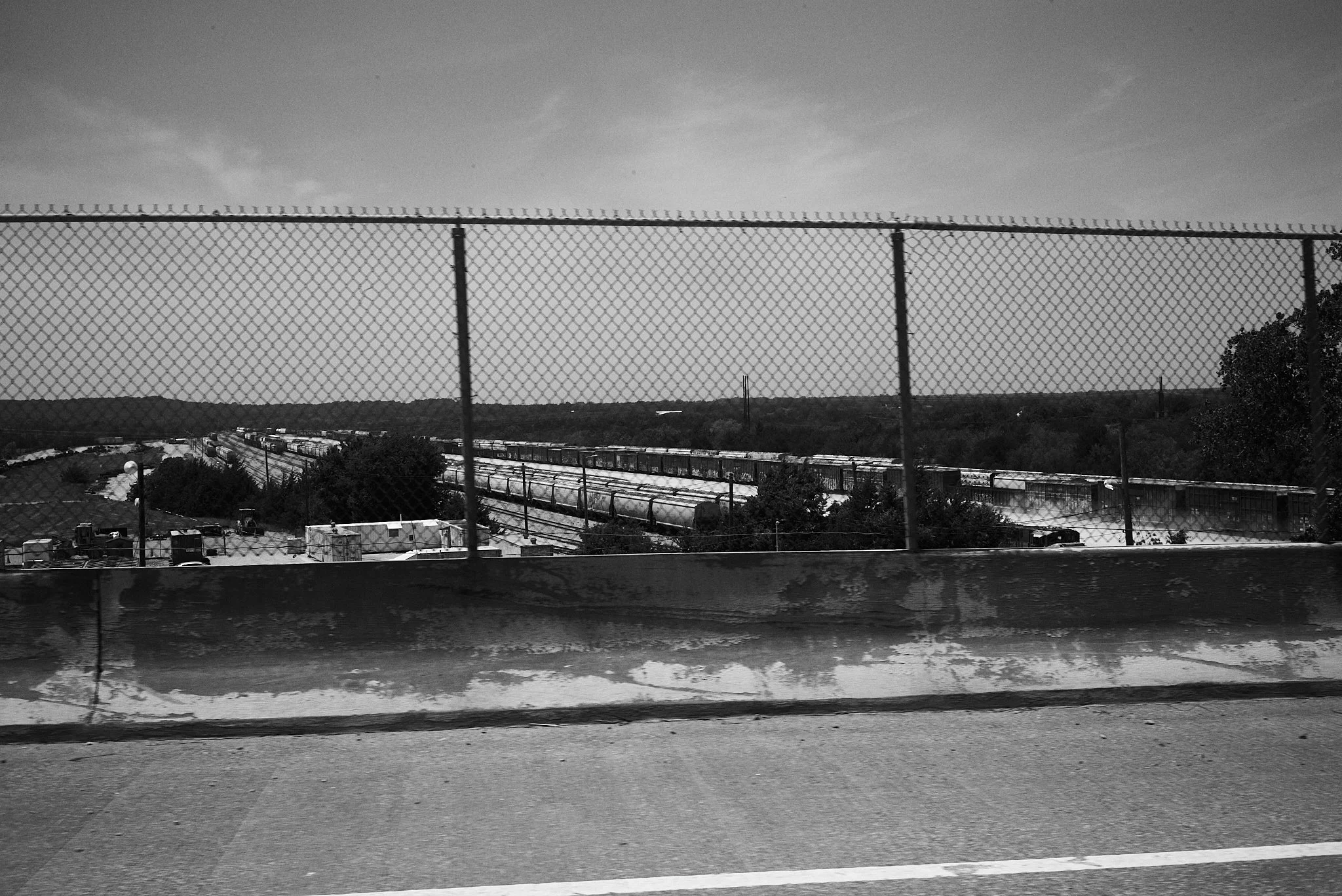 Black and white photo of a train on railway tracks behind a chain-link fence, with hills and trees in the background.