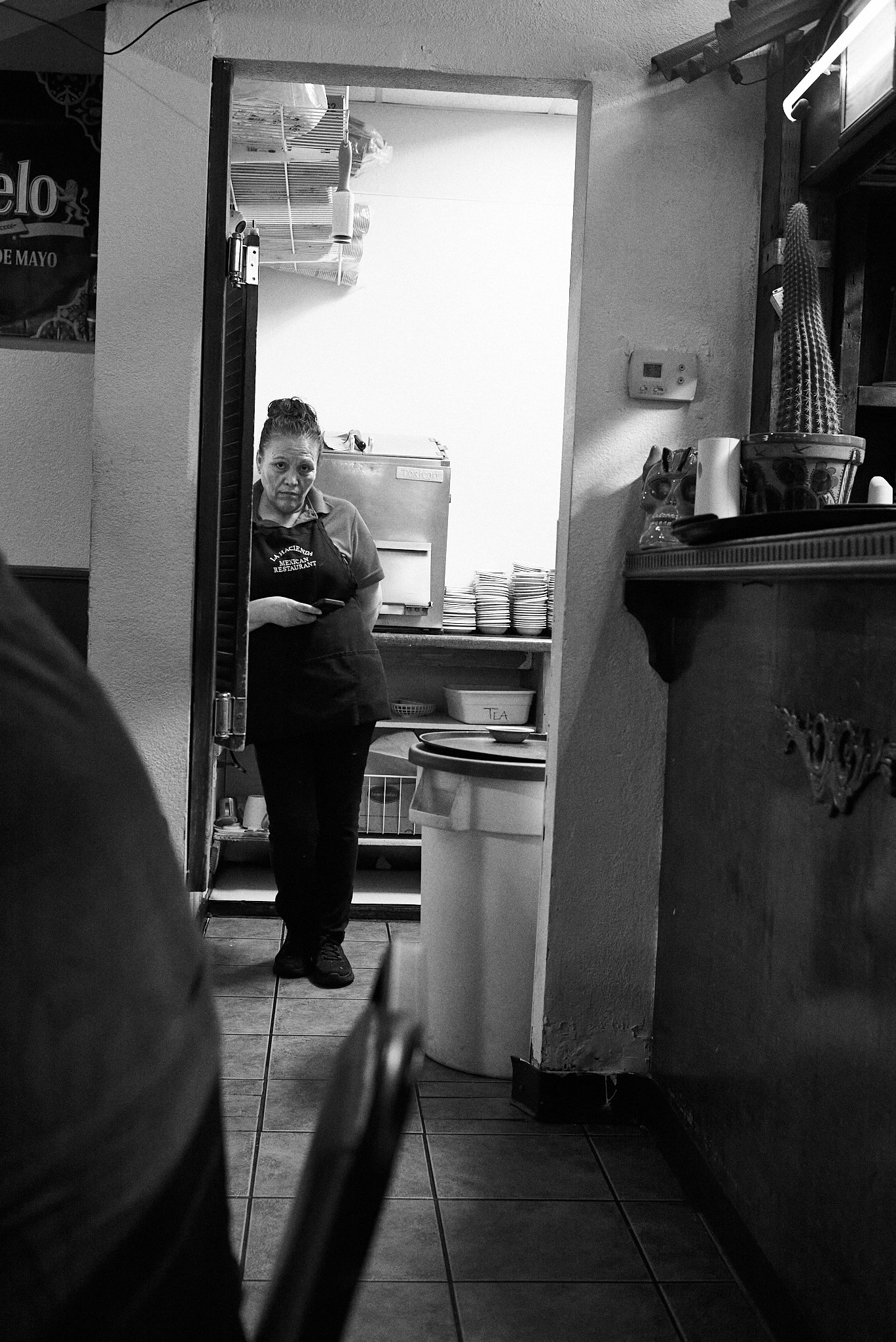 A woman in a restaurant or kitchen stands in a doorway looking towards the camera, holding a phone. The room behind her has dishes stacked on a shelf, a tray with bowls labeled 'Tea,' and a small refrigerator.