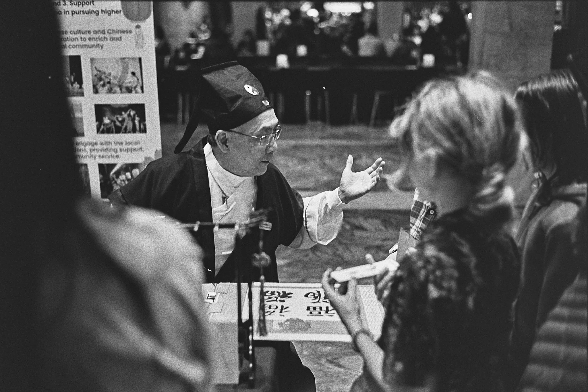 A person dressed in traditional Asian attire, possibly a guru or instructor, with glasses and a headpiece, is speaking to three women, one with light-colored hair holding a piece of paper. The scene takes place indoors with other people and a sign in