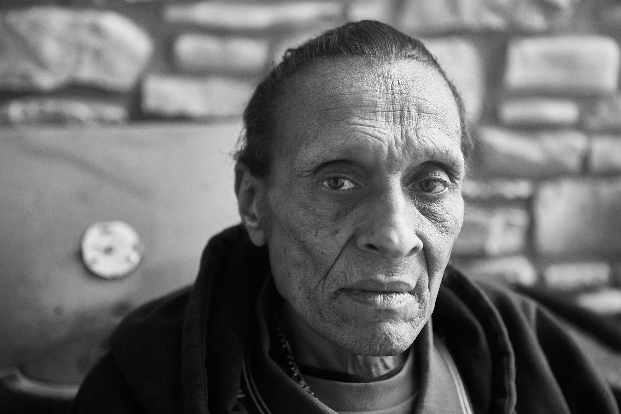 Black and white close-up photograph of an elderly man with a serious expression, dark hair pulled back, wearing a dark jacket, with a brick wall in the background.
