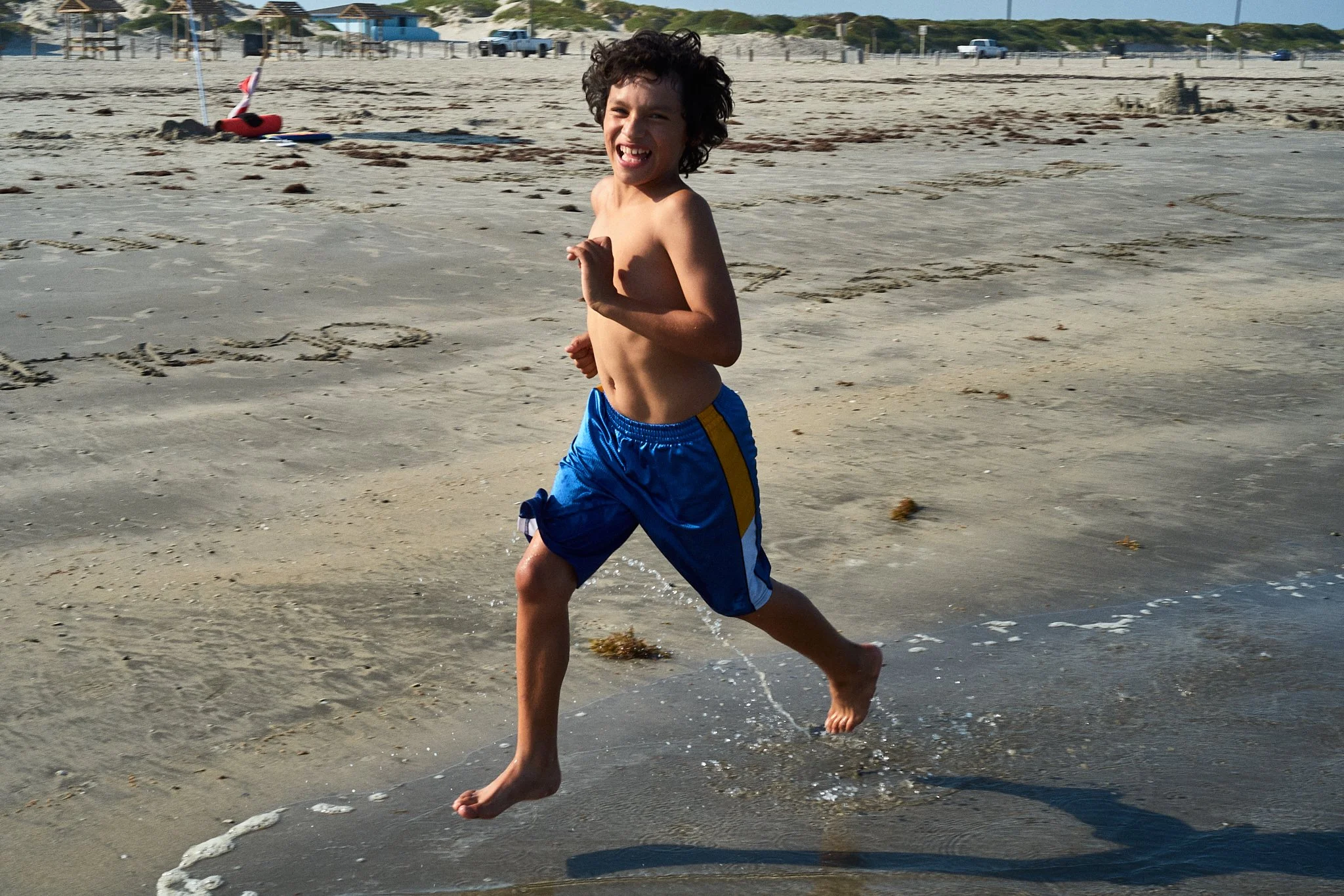 A young boy with curly dark hair, shirtless, wearing blue swimming shorts, running and playing at the beach in shallow water, smiling and looking happy.