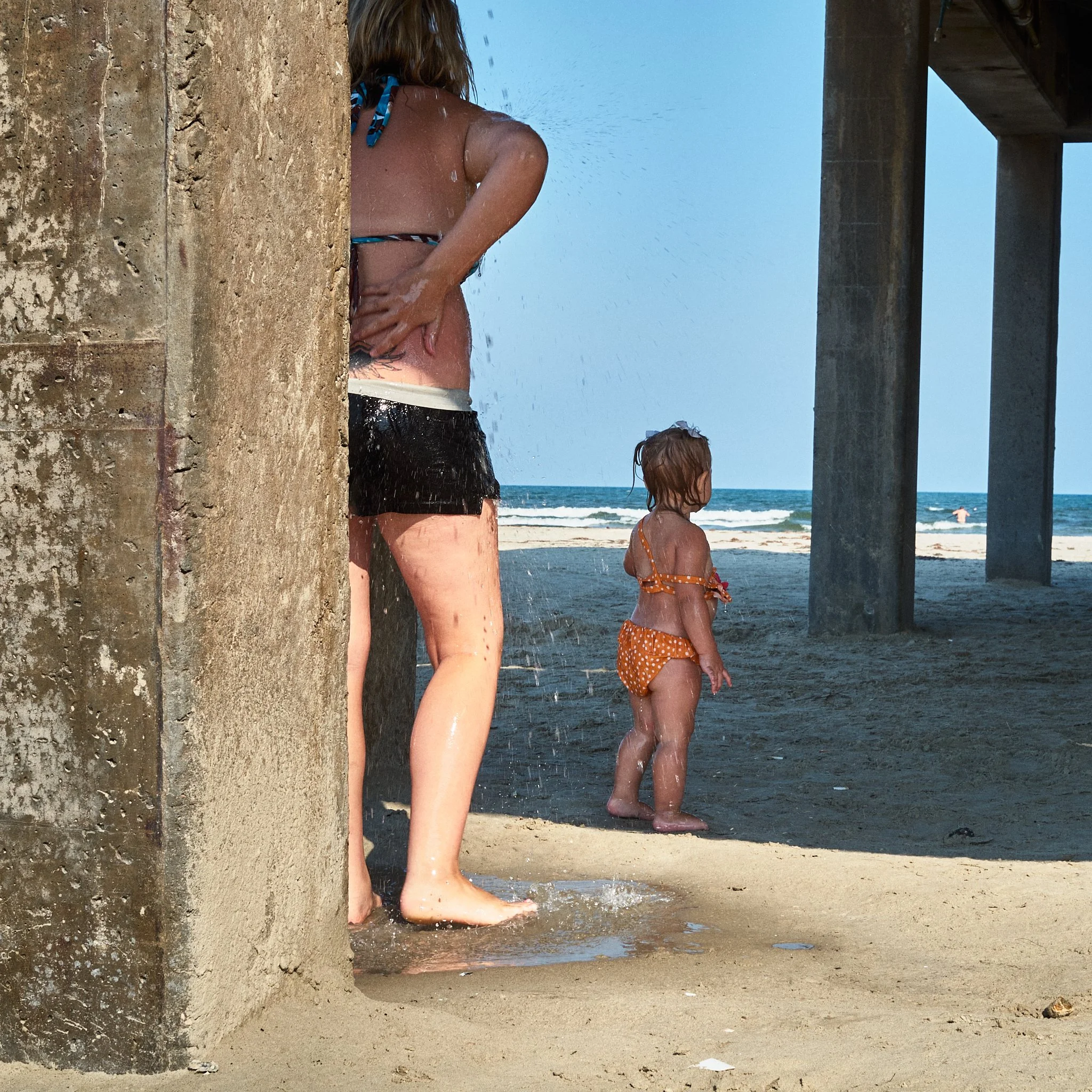 A woman and a small girl standing under a pier at the beach, with ocean waves in the background. The woman is washing her hair under a concrete pillar, and the girl is looking towards the ocean.