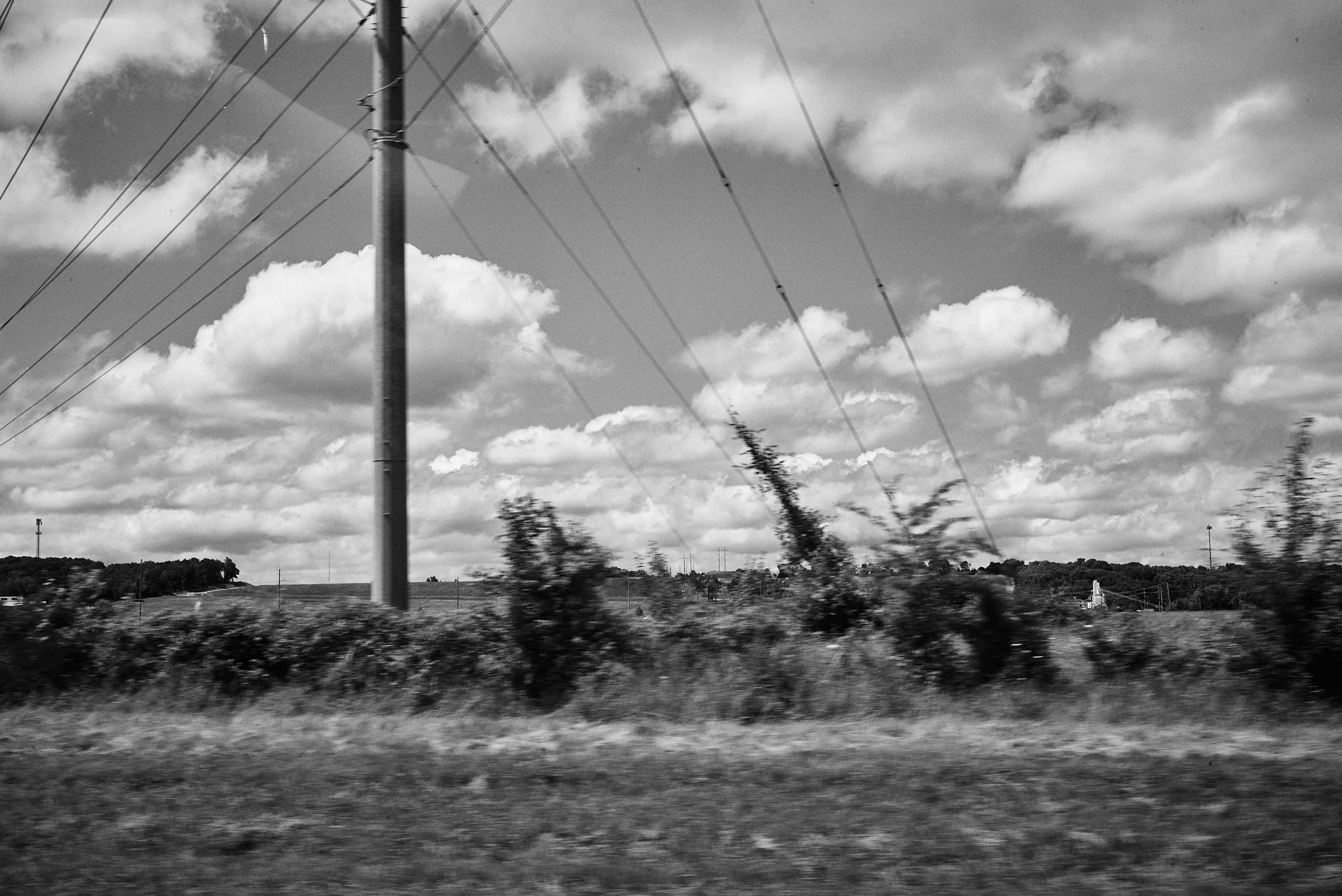 Black and white photograph of a rural landscape with power lines and poles, clouds in the sky, and trees in the distance.